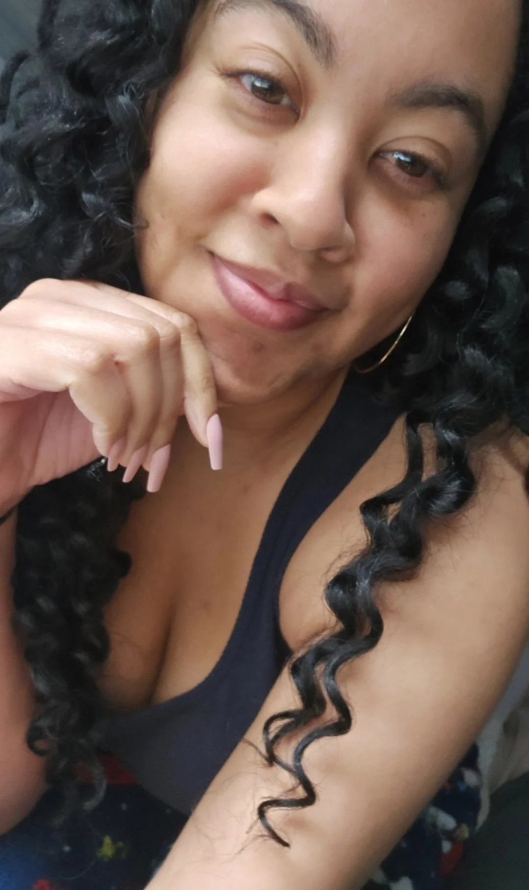 A woman with long, curly black hair, wearing a black tank top and gold hoop earrings, resting her chin on her hand with pink acrylic nails, smiling softly.
