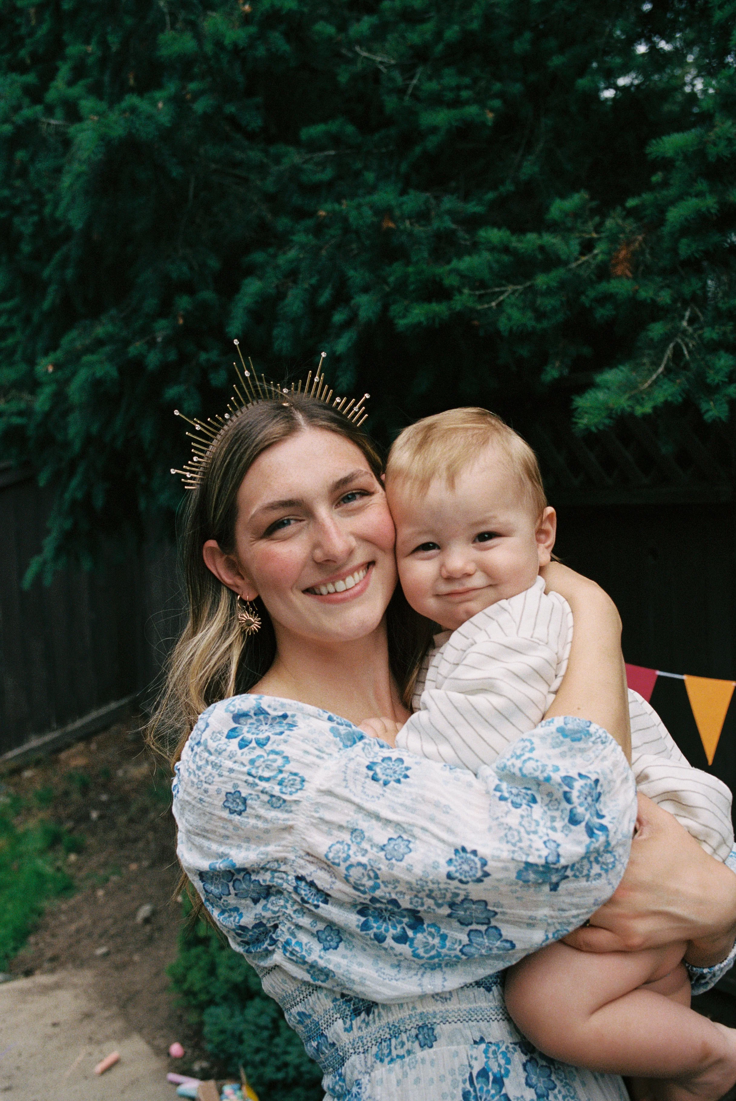 A woman with long brown hair, wearing a blue and white floral dress and a golden crown, is smiling and holding a child in her arms. They are outdoors with green trees and a black fence in the background.