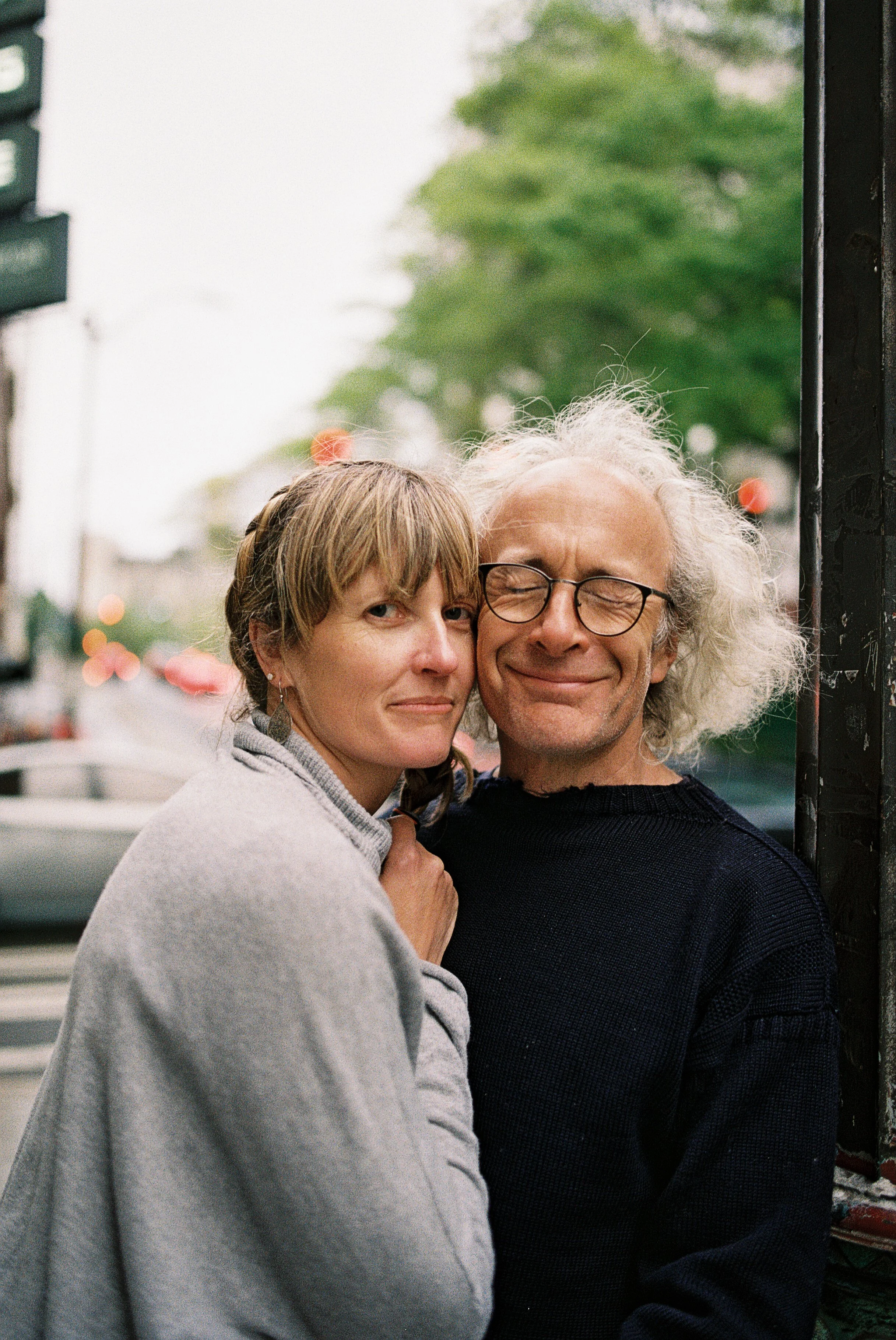 A woman with light brown hair and a man with white, curly hair and glasses stand close together outdoors, smiling with their eyes closed. The woman wears a gray hoodie, and the man wears a dark sweater. Blurred trees and city street elements are in the background.