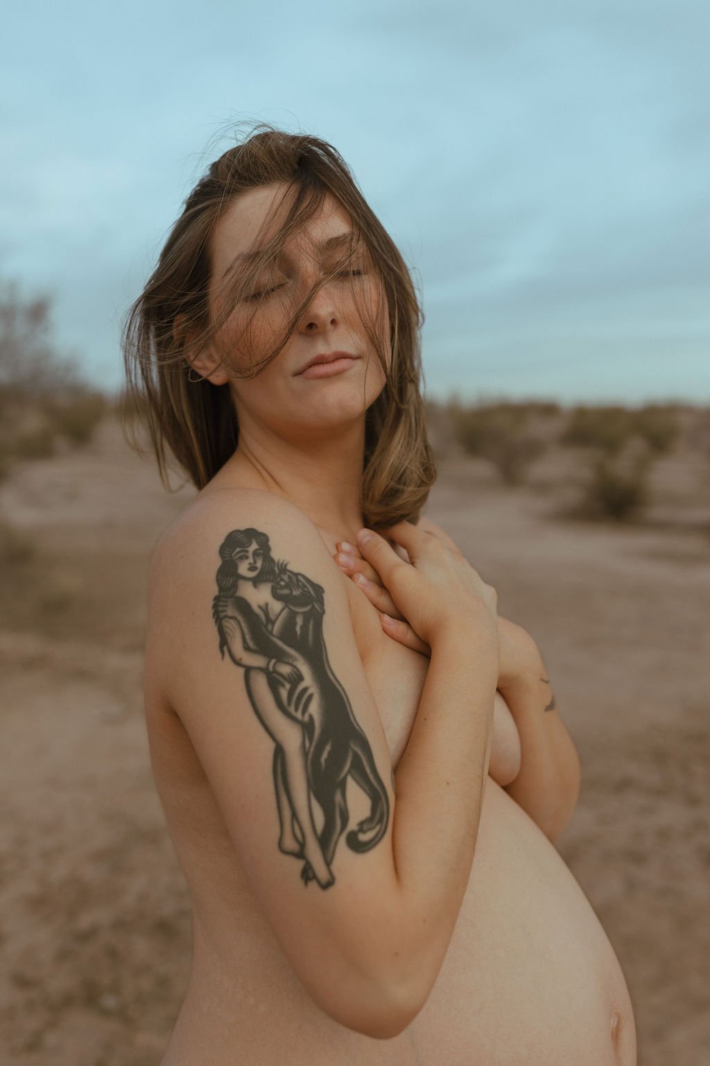 A woman with medium-length brown hair and a tattoo on her left arm, standing in a desert with sparse bushes, her eyes closed and her hands resting on her chest.