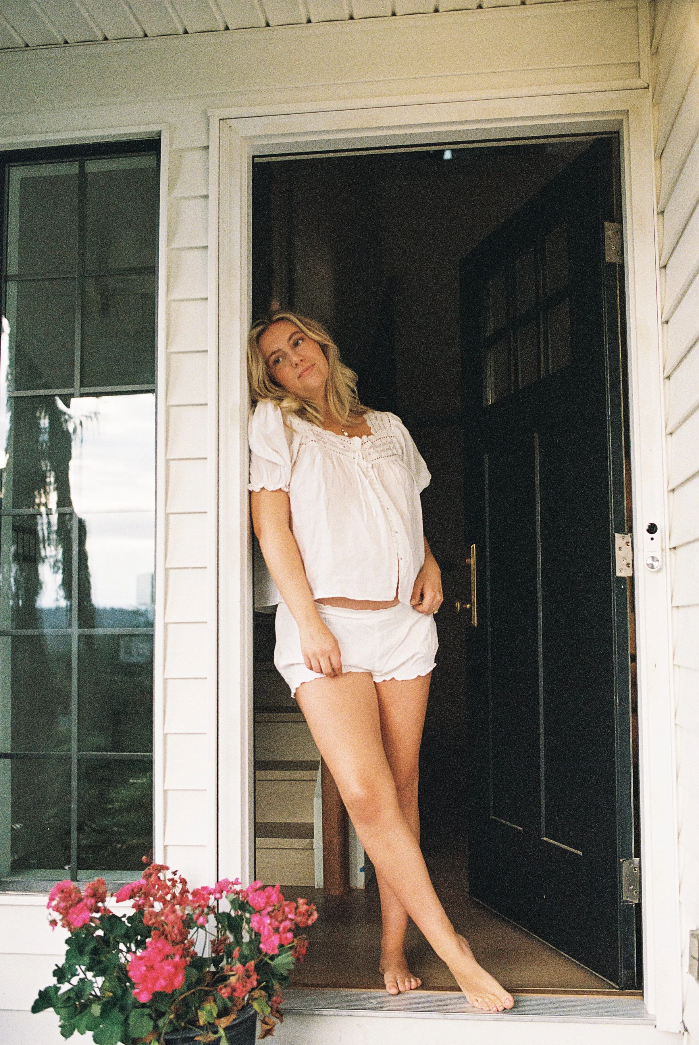 A pregnant woman stands barefoot in a doorway, wearing a white, loose blouse and matching shorts, with pink flowers in the foreground underneath a window. She looks off to the side with a relaxed expression.