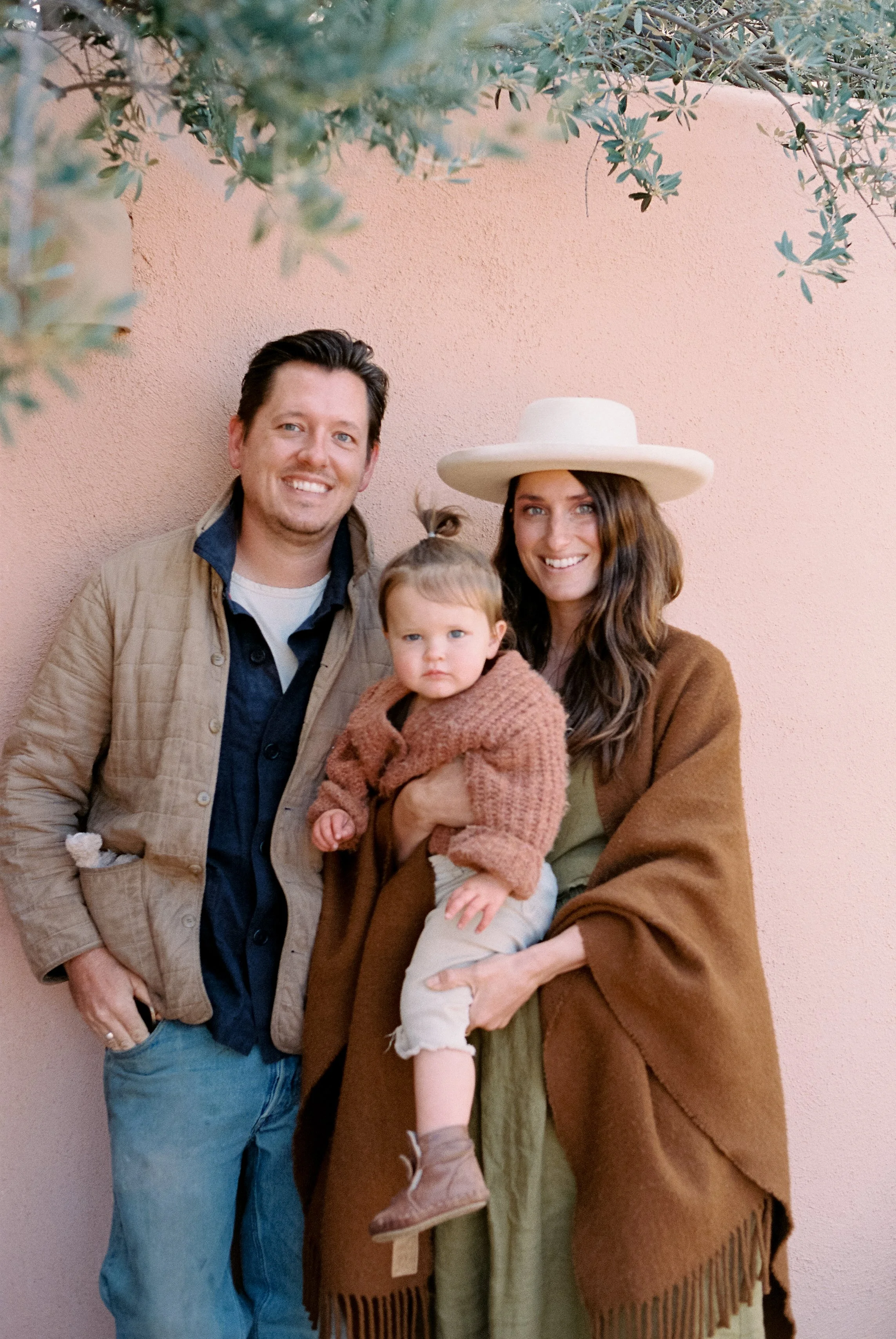 A family of three standing against a pink wall for a portrait.