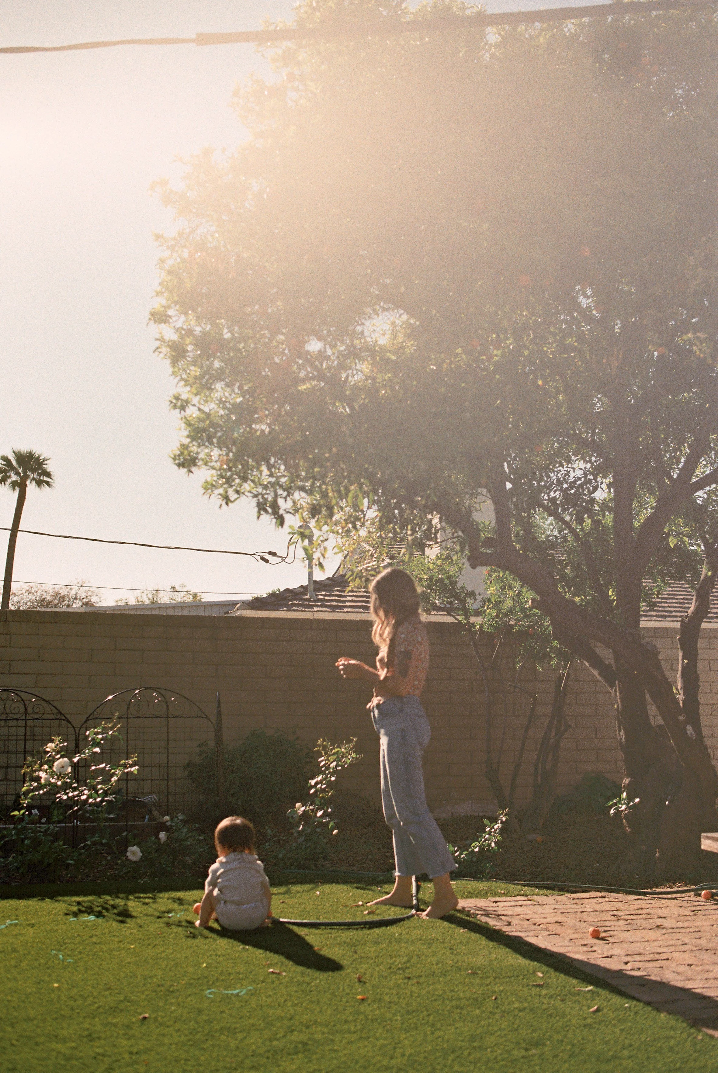A woman and two children playing in a backyard on a sunny day. The woman is standing near a brick pathway, with one child sitting on the grass and the other standing nearby. There are trees, a garden fence, and a house with a tiled roof in the backgr