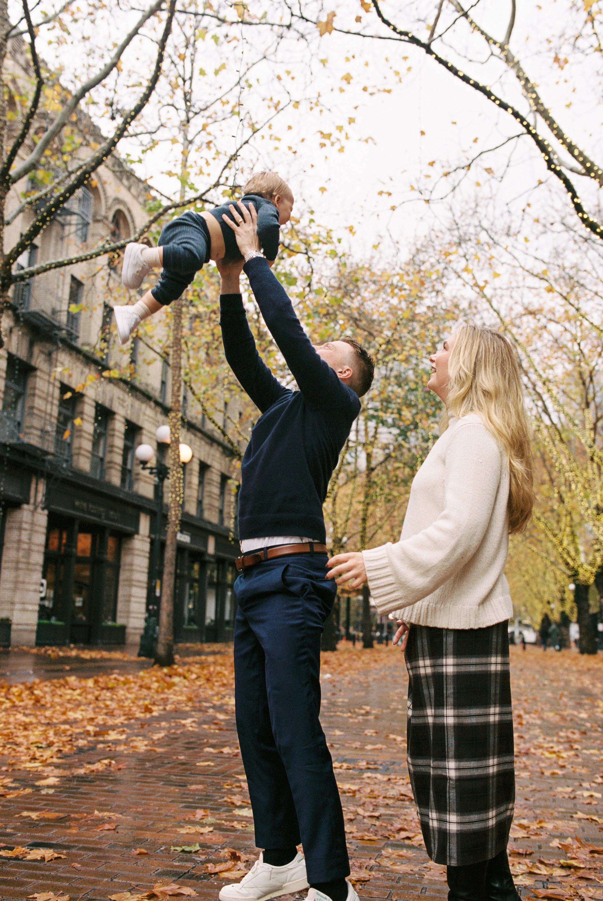 A man lifting a young boy in the air while a woman watches. They are outside on a rainy autumn day with fallen leaves on the ground and tree branches with lights hanging on them.
