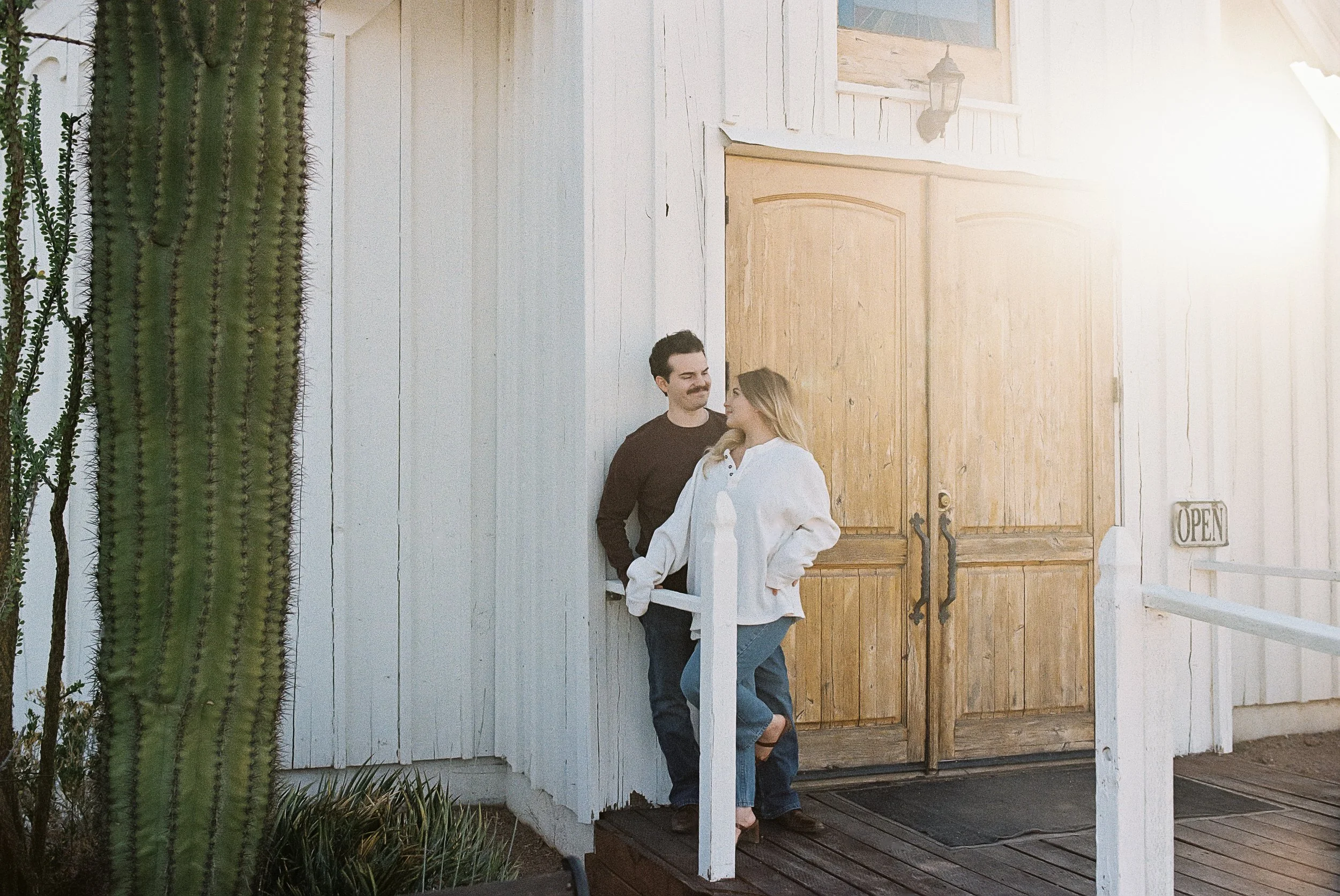 A man and woman standing near a wooden door of a small white building with a sign that says 'OPEN' on the side, a large cactus on the left side, and sunlight on the right side.