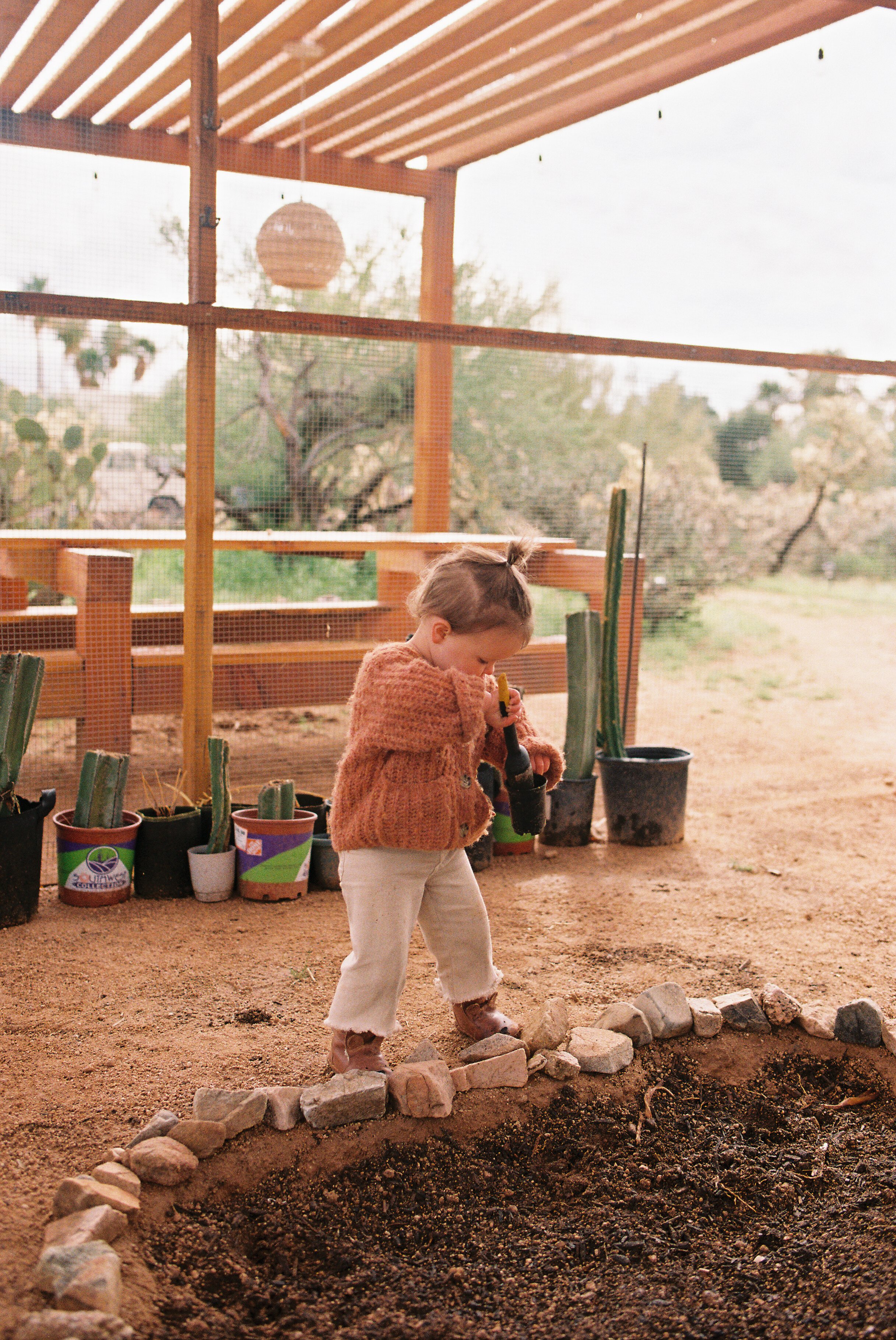 A child gardening in their home garden. There are potted plants behind her and a wooden structure with a hanging lantern in the background.