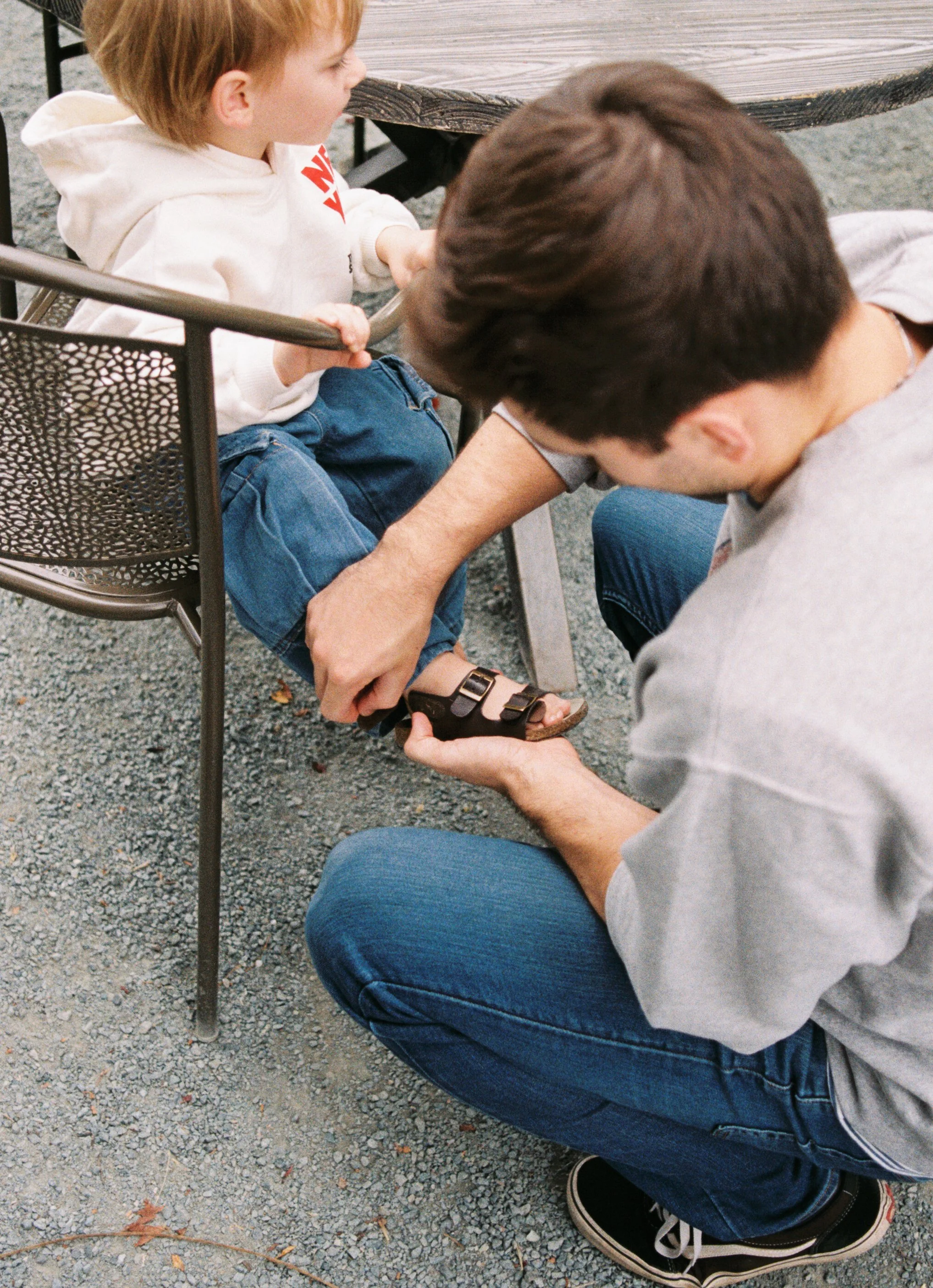 A man kneeling on gravel ground tying the sandal of a young boy sitting in a metal chair outdoors.