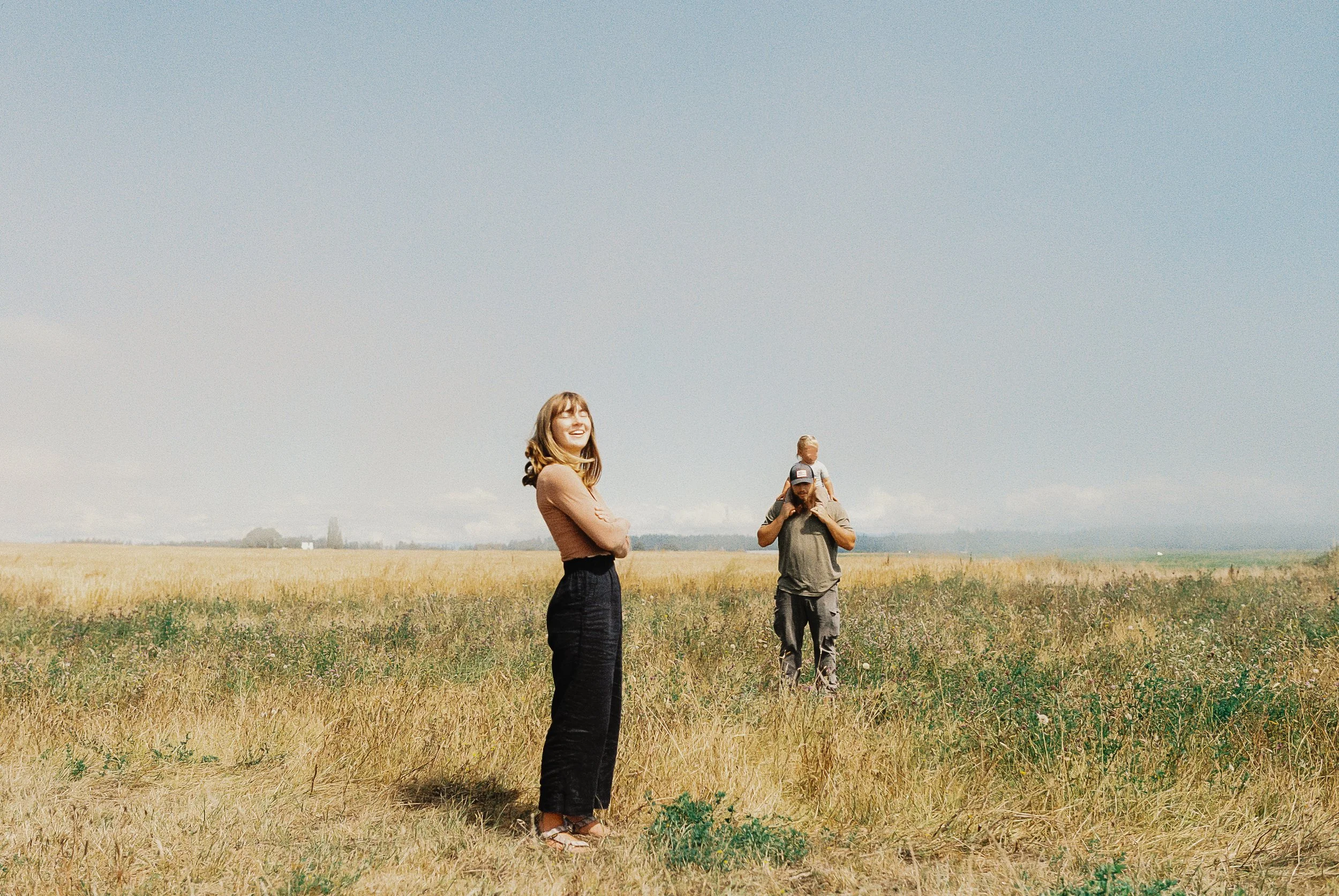 Three people standing in a field under a clear sky, with mountains visible in the distance. One woman in the foreground is smiling, wearing a brown top and black skirt. Behind her, a man is carrying a child.