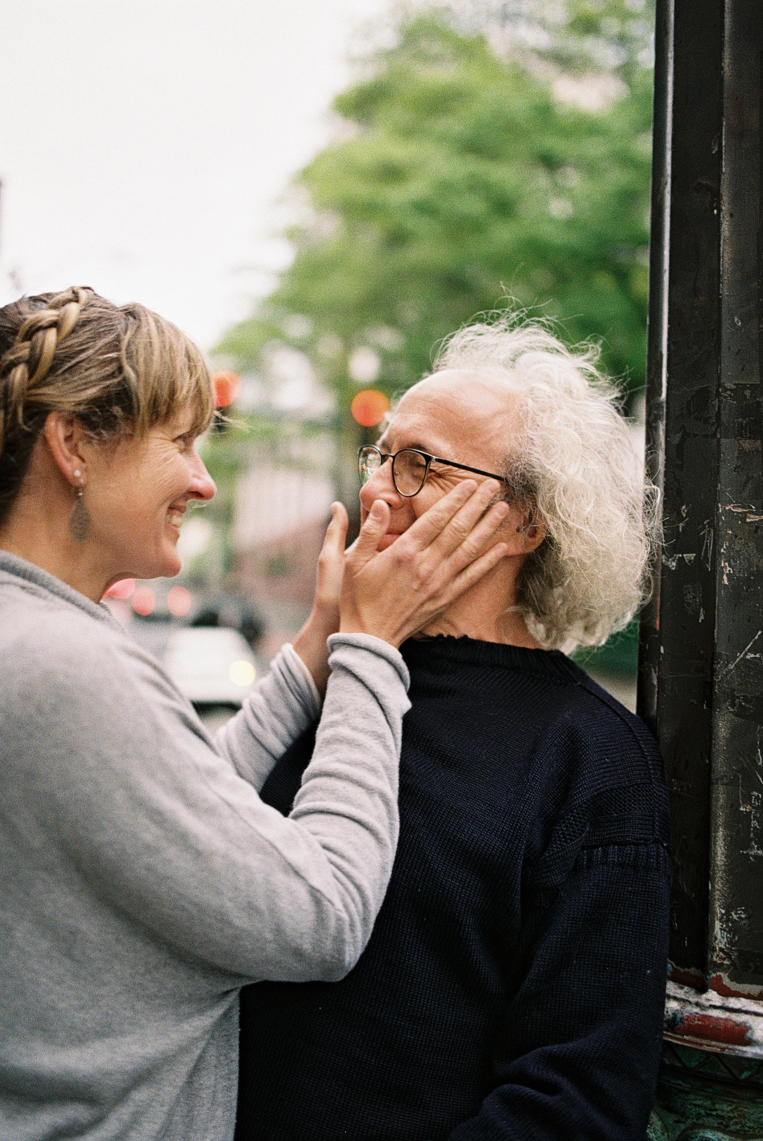 A couple stands on a street corner and they both smile fondly at each other. The background is blurred with green trees and city lights.