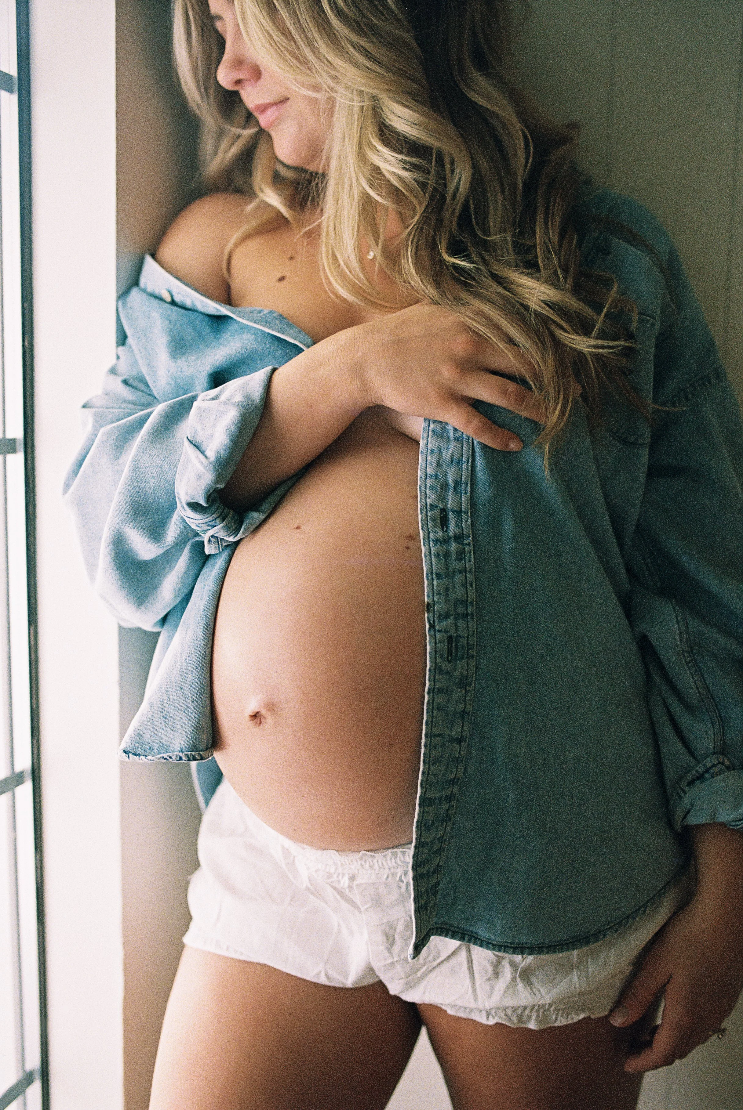 A woman with blonde hair, leaning against a wall, posing for a maternity photo including her belly.