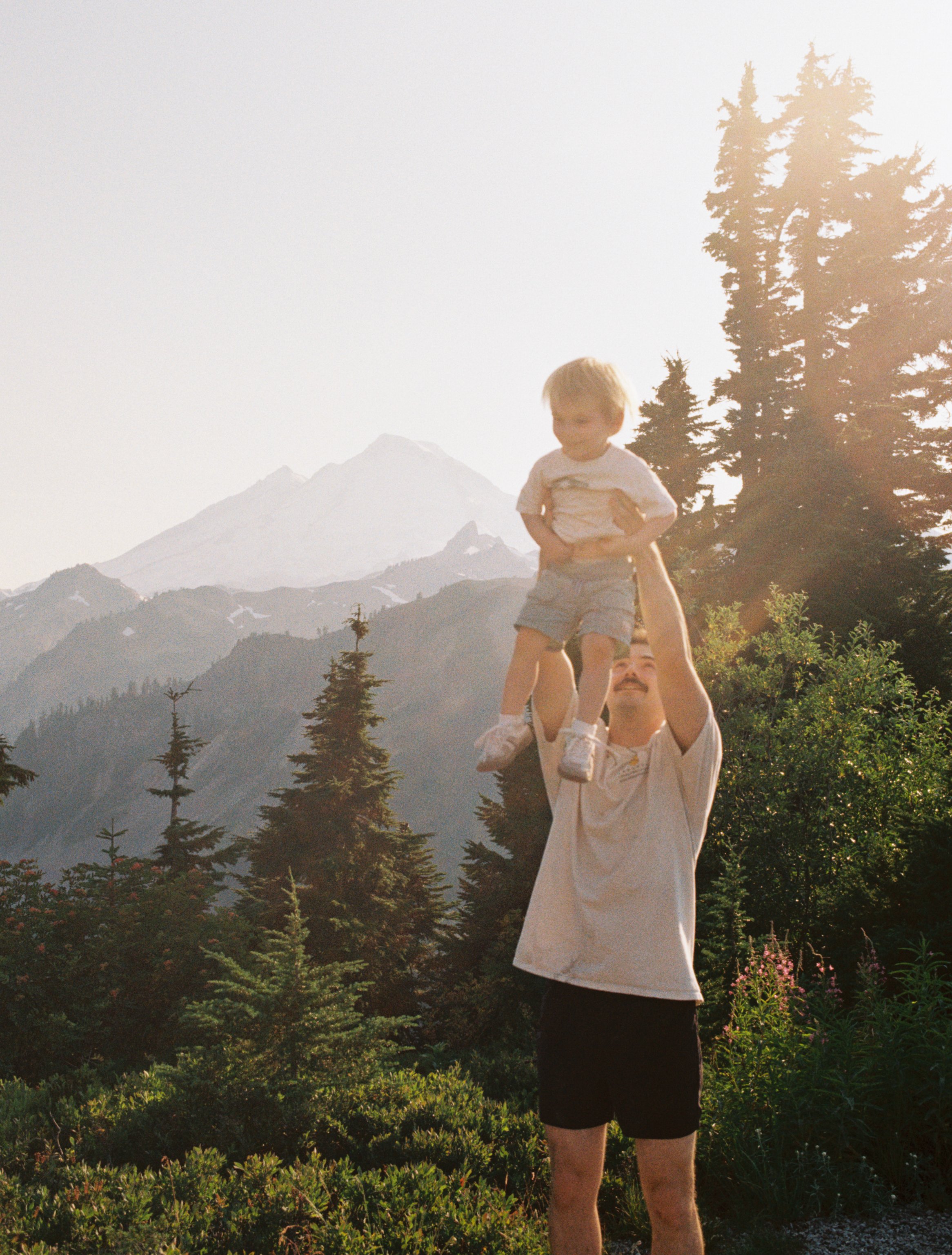 A man lifting a young boy in the air in a mountainous outdoor setting with trees and snow-capped peaks in the background during sunset.