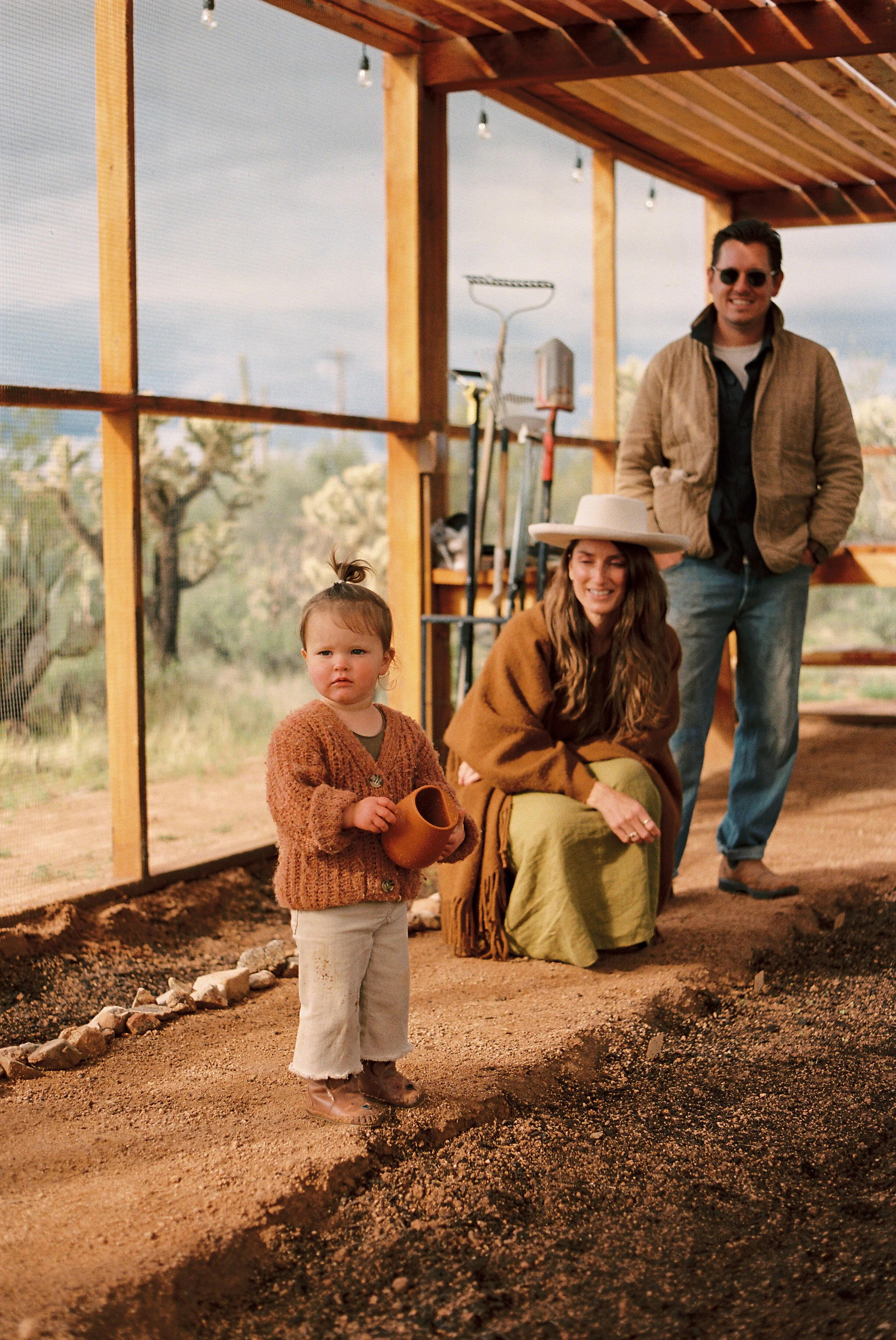 A young girl holding a small terra cotta pot, standing on a dirt construction site with a partially built wooden structure and two adults in the background.