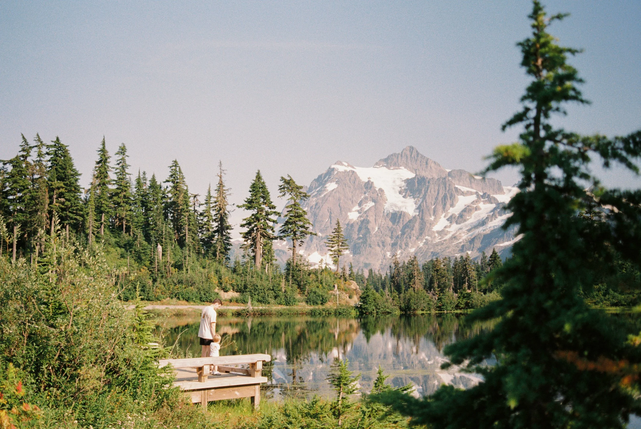 A serene lake surrounded by tall evergreen trees with snow-capped mountains in the background. There is a person with a child standing on a small wooden dock near the water.