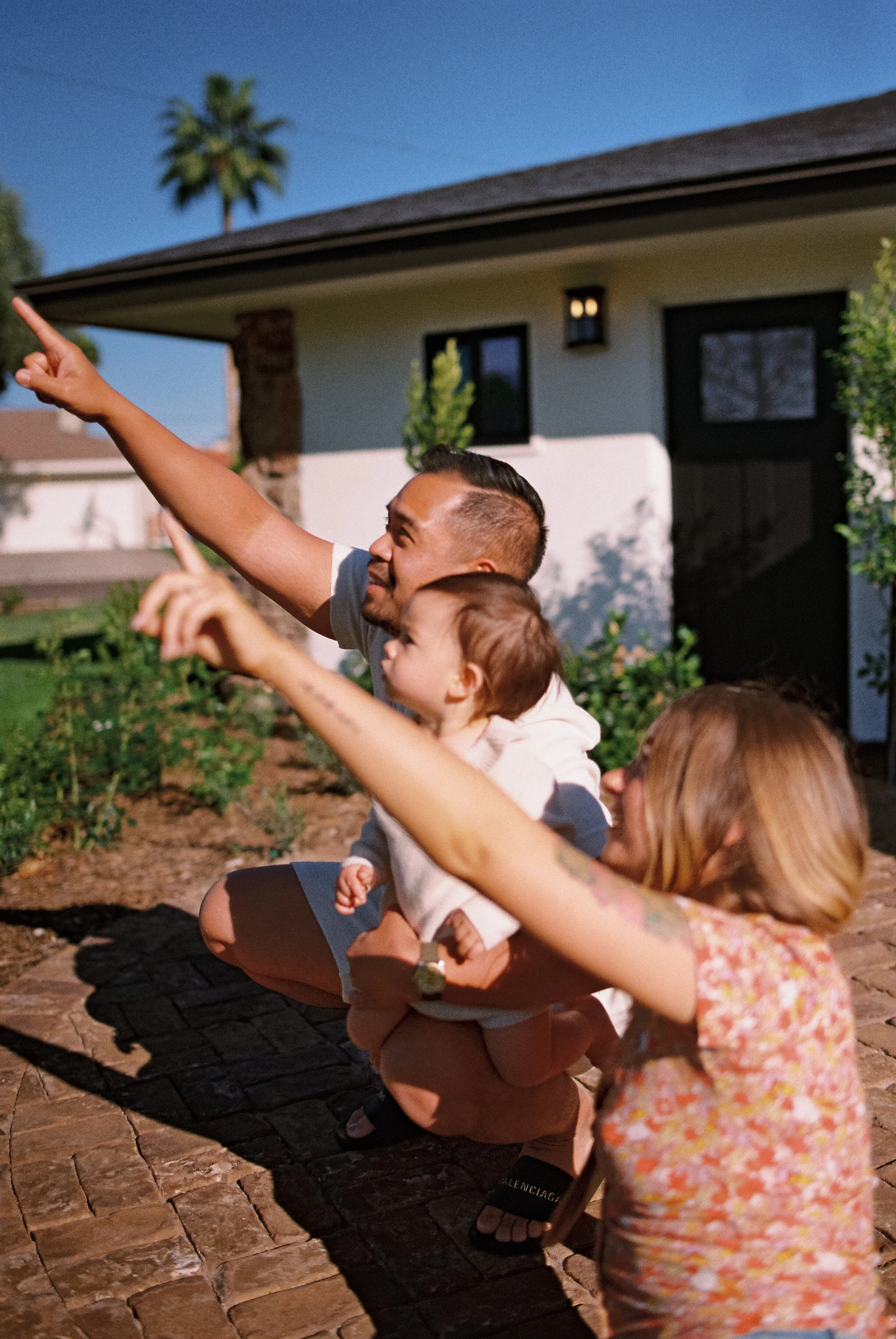 A group of people, including children and an adult with short dark hair, are crouching on a brick pathway outside a house, pointing at something in the sky during daylight.