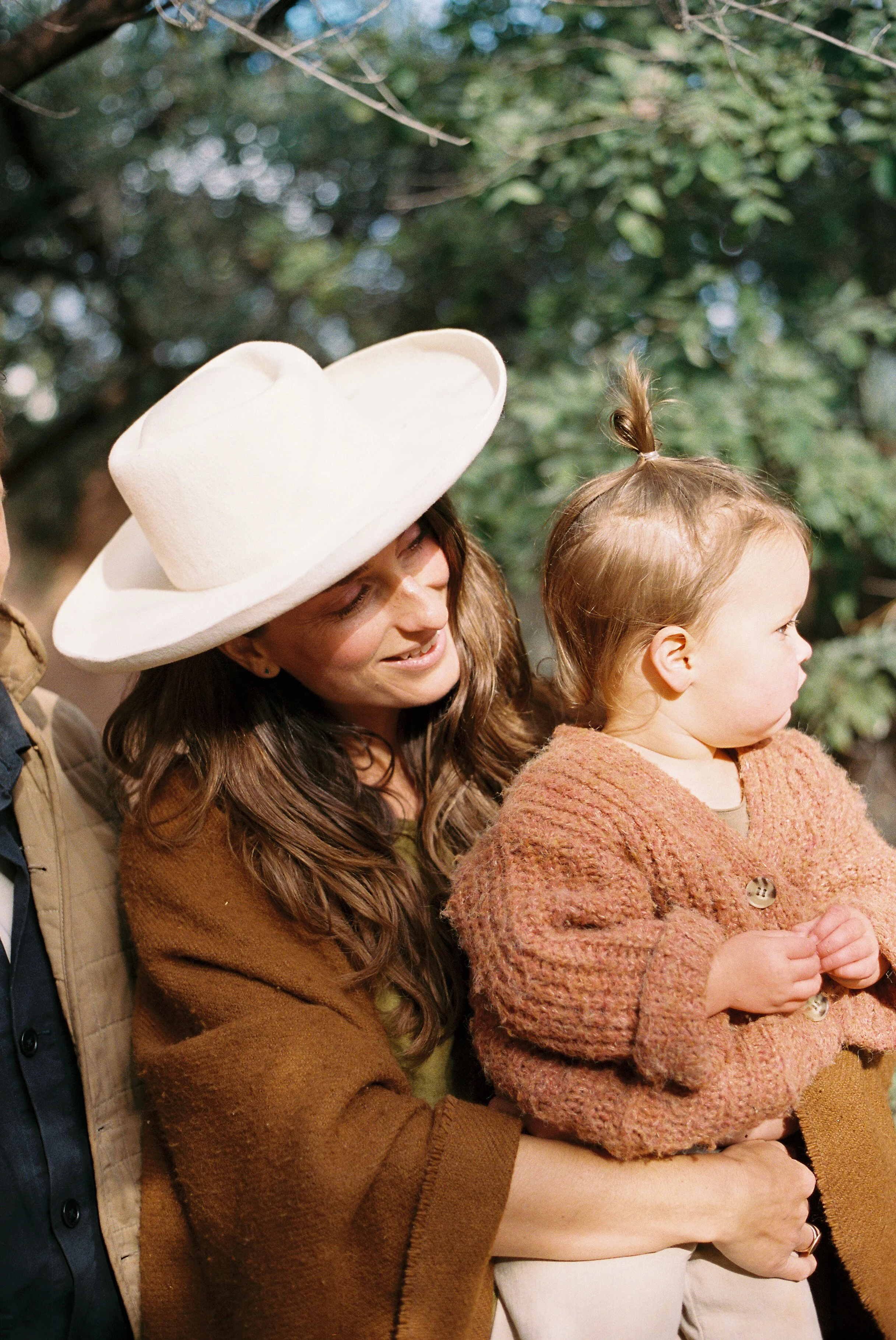 A woman with long brown hair wearing a white hat and brown sweater holding a young girl with blonde hair in a sweater outdoors, with green trees in the background.