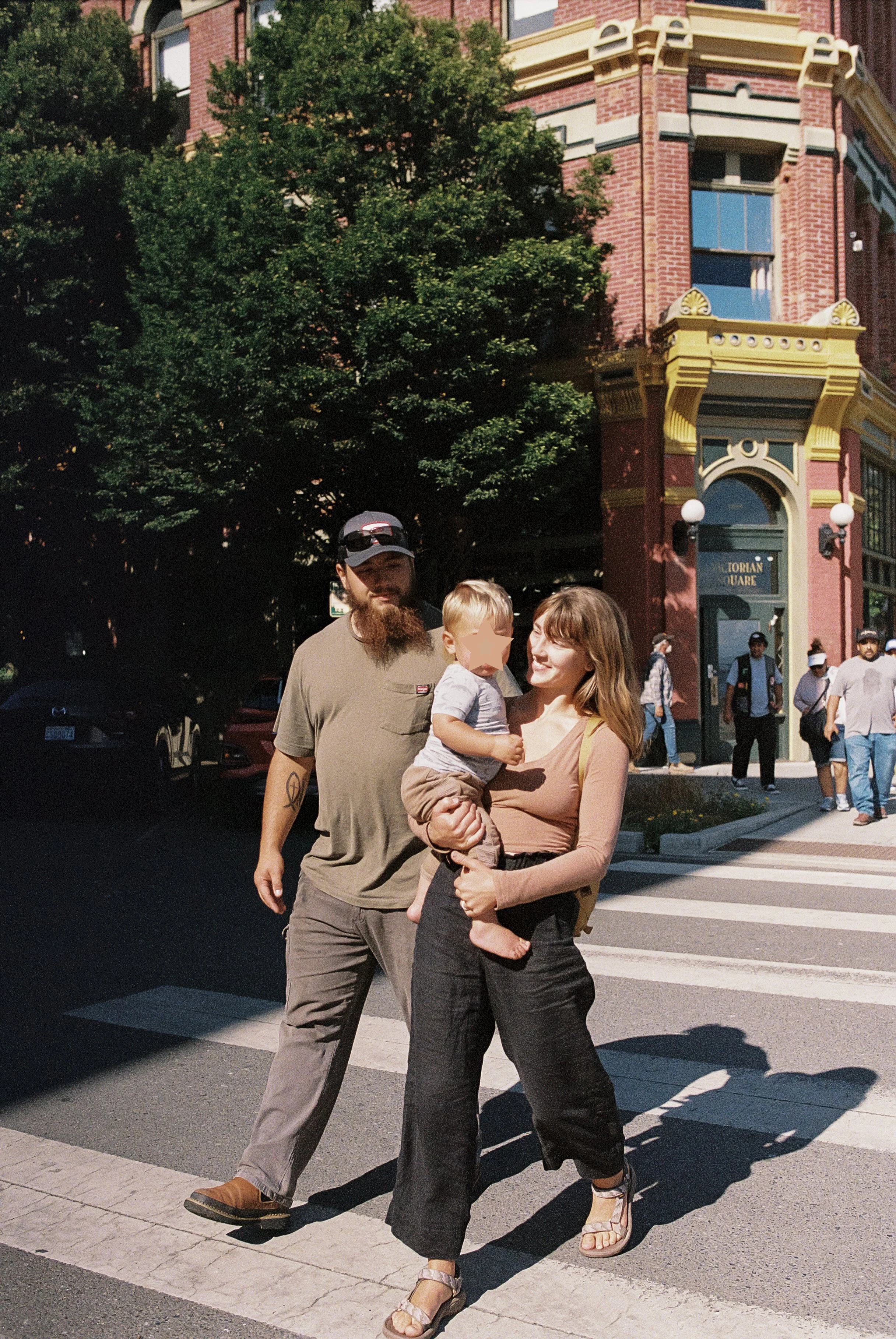 A family of three, a man, a woman, and a young boy, walking across a crosswalk in front of a red brick building with ornate yellow and gray architectural details. The woman is holding the child, and the man is walking beside them. There are pedestria