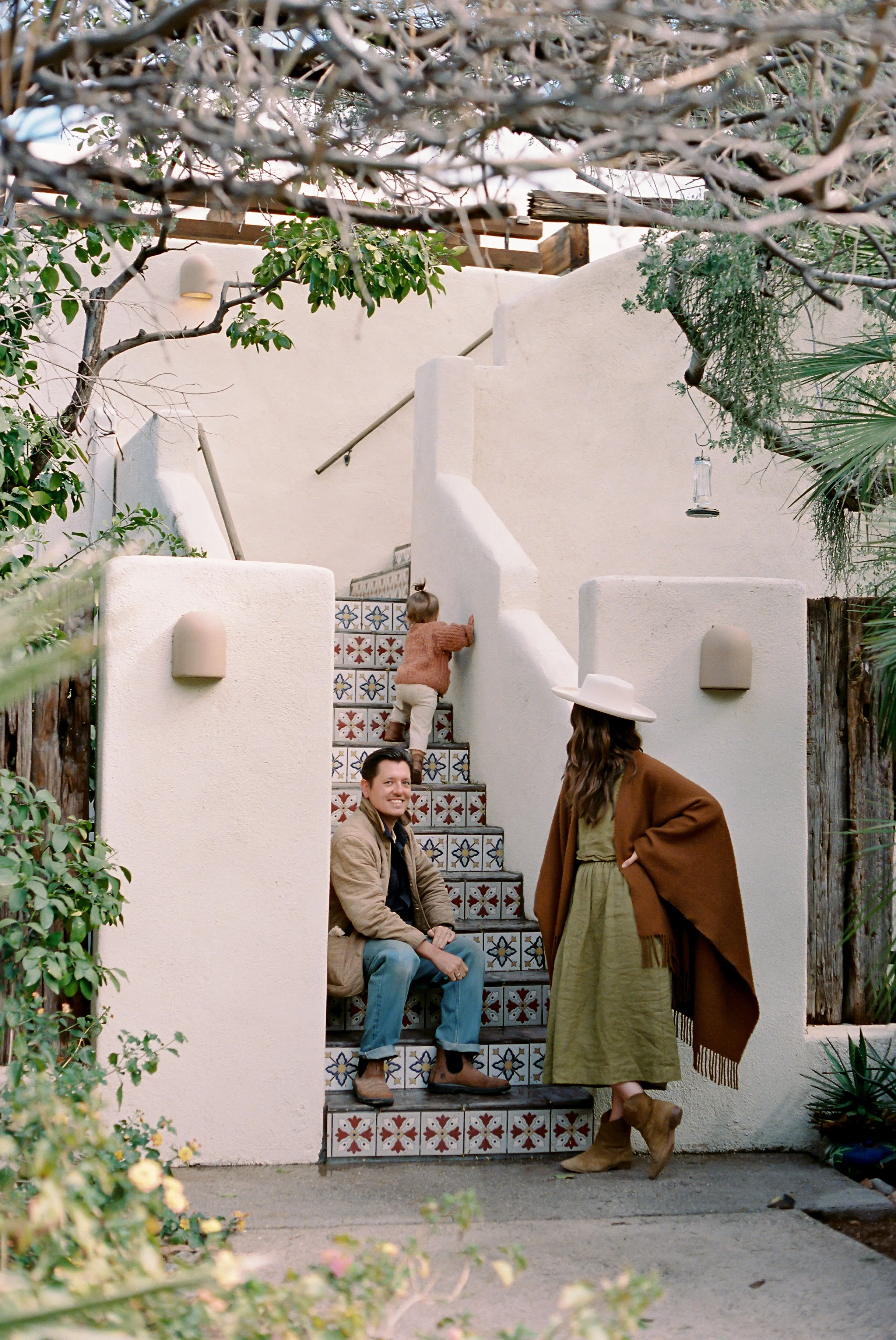 A family poses on a staircase. The staircase has colorful decorative tiles on the risers, and is surrounded by greenery, including a tree with branches overhead and plants along the sides.