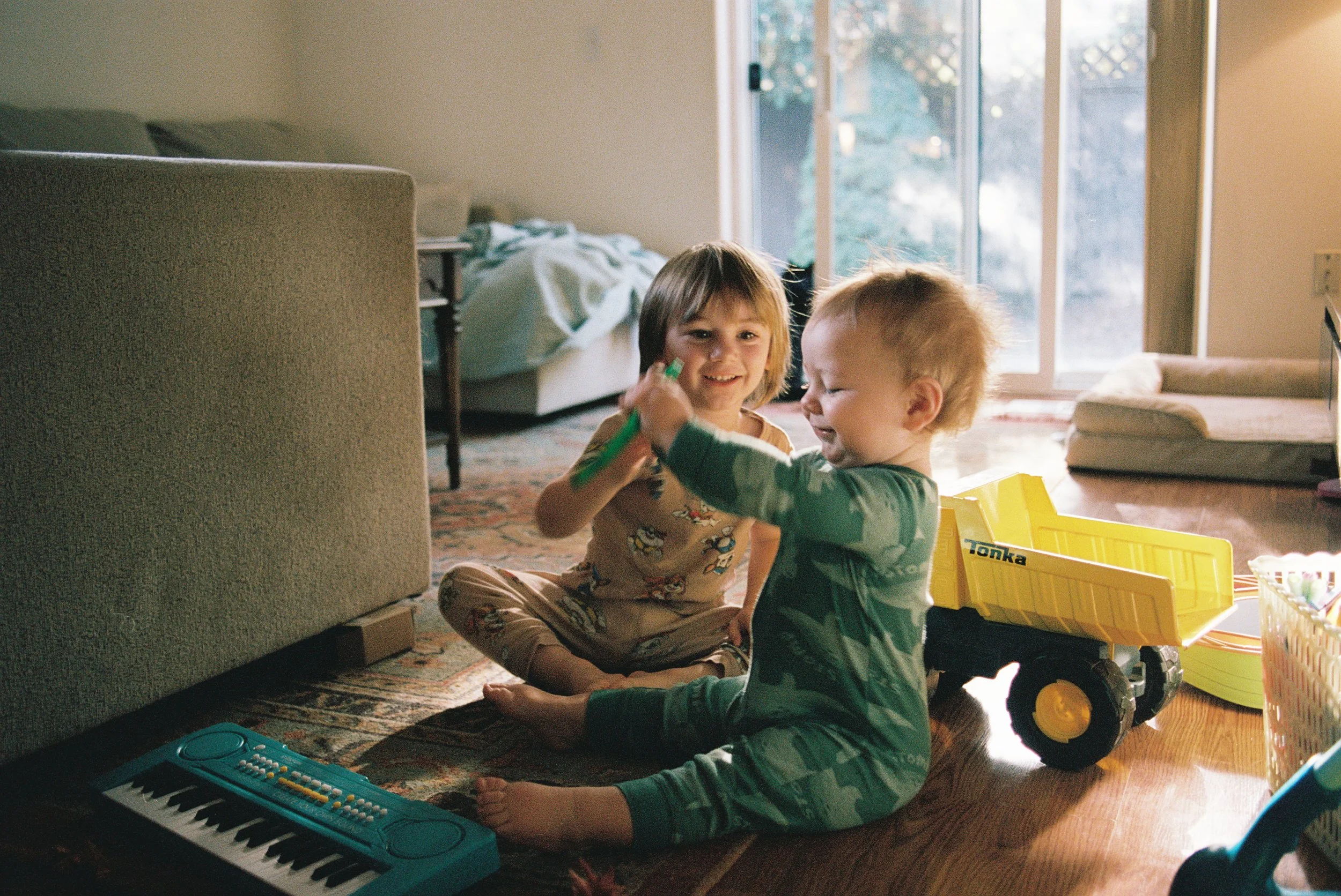 Two young children, a boy and a girl, playing with toys on the floor of a sunlit living room. Large glass sliding doors in the background let in natural light, showing a backyard scene.