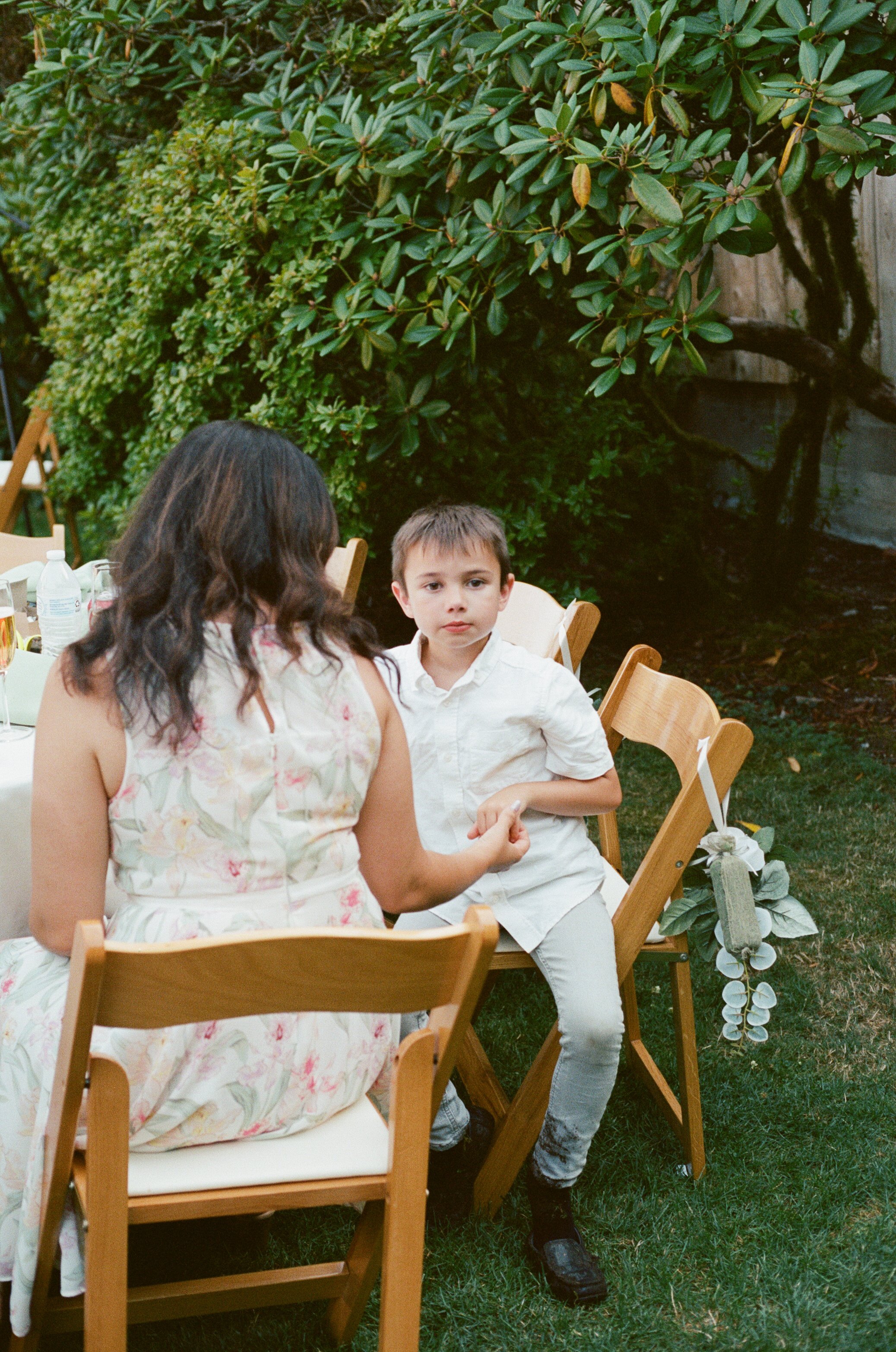 A young boy in white shirt and gray pants is sitting on a wooden chair outdoors at a gathering, listening to a woman in a floral dress who is speaking. There are other chairs, a table with drinks, and lush greenery with a large tree in the background