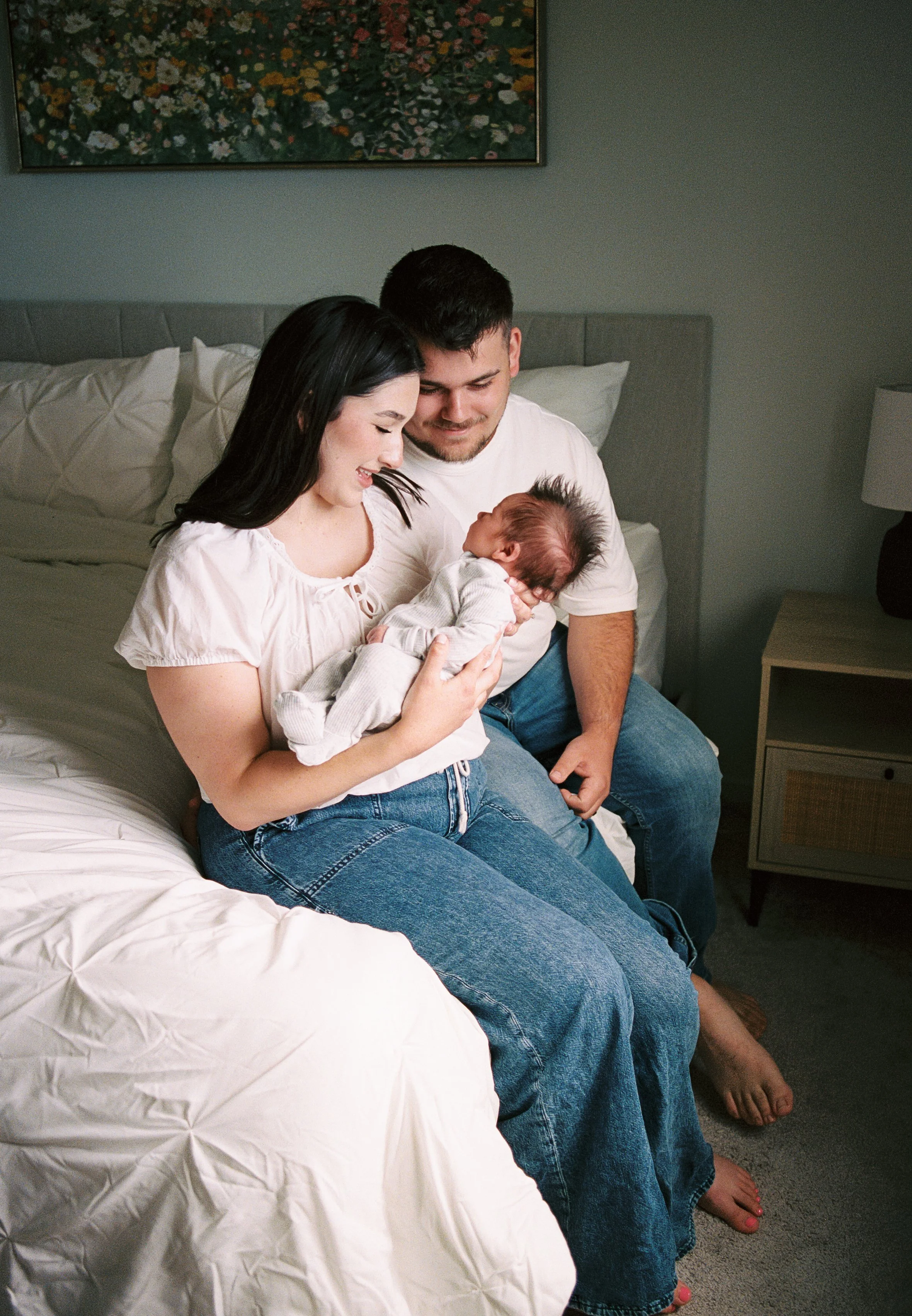 A family of three sitting on a bed in a bedroom, smiling and cuddling a newborn baby. The woman has black hair and is wearing a white shirt, and the man has short dark hair and is wearing a white t-shirt and blue jeans. The baby is wrapped in a light