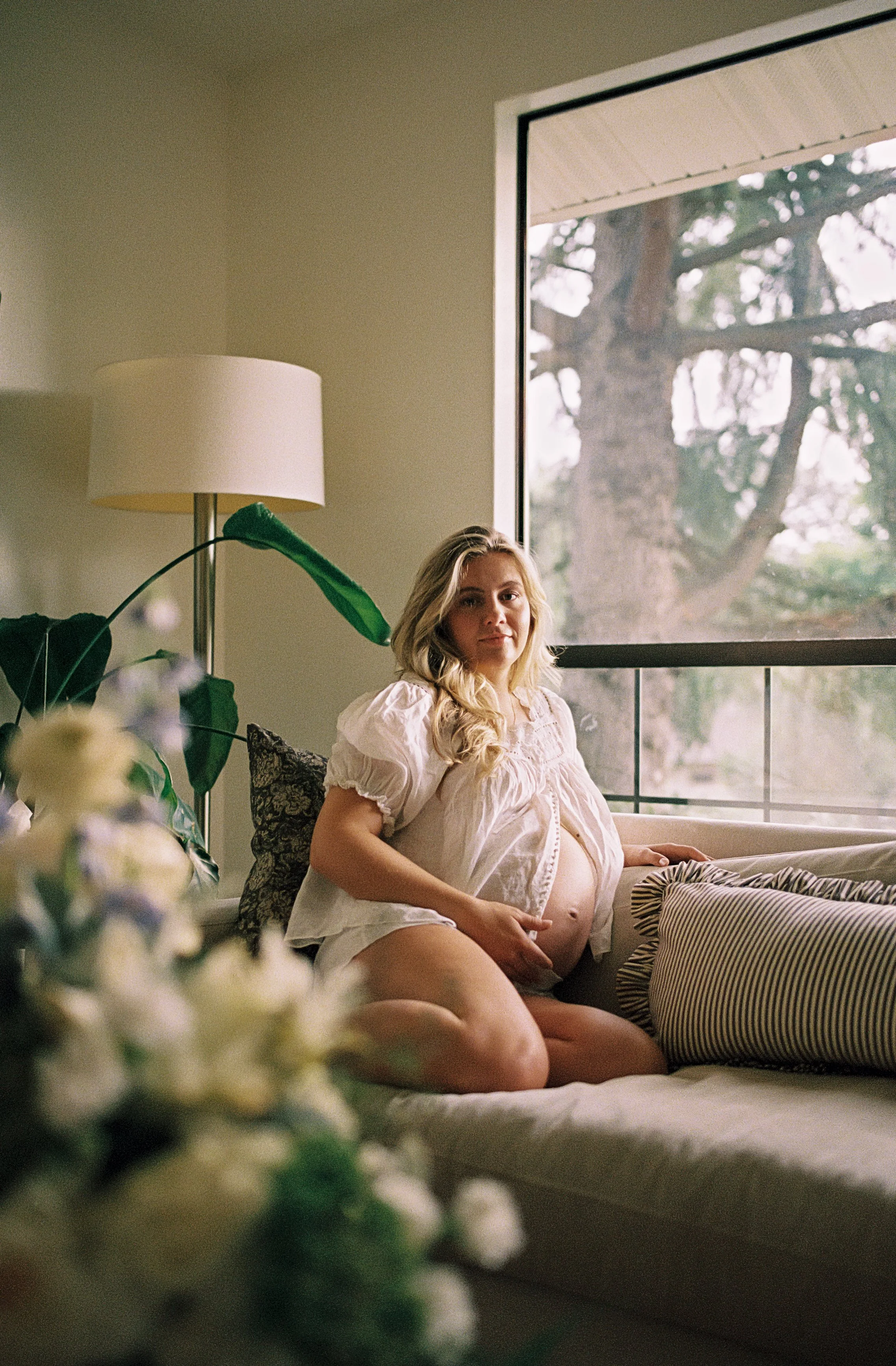 A pregnant woman with long blonde hair sitting on a sofa in a living room, looking at the camera. She is wearing a white blouse, and her hand is resting on her belly. The room has a large window showing trees outside, and a tall floor lamp with a white shade behind her. There are various cushions on the sofa, and flowers are in the foreground.