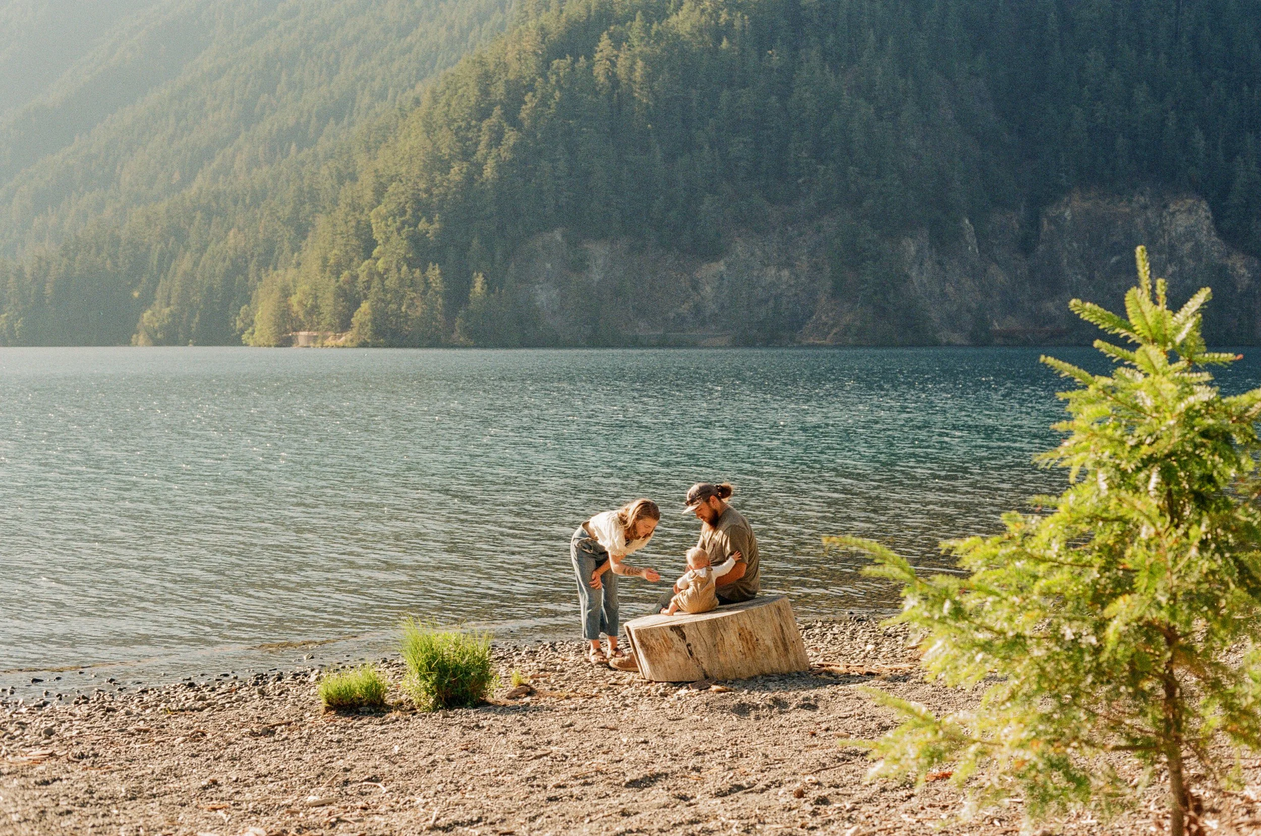 A family of three is on a rocky beach by a lake. In the background, there are forested mountains and a lake under a clear sky, with sunlight reflecting off the water.