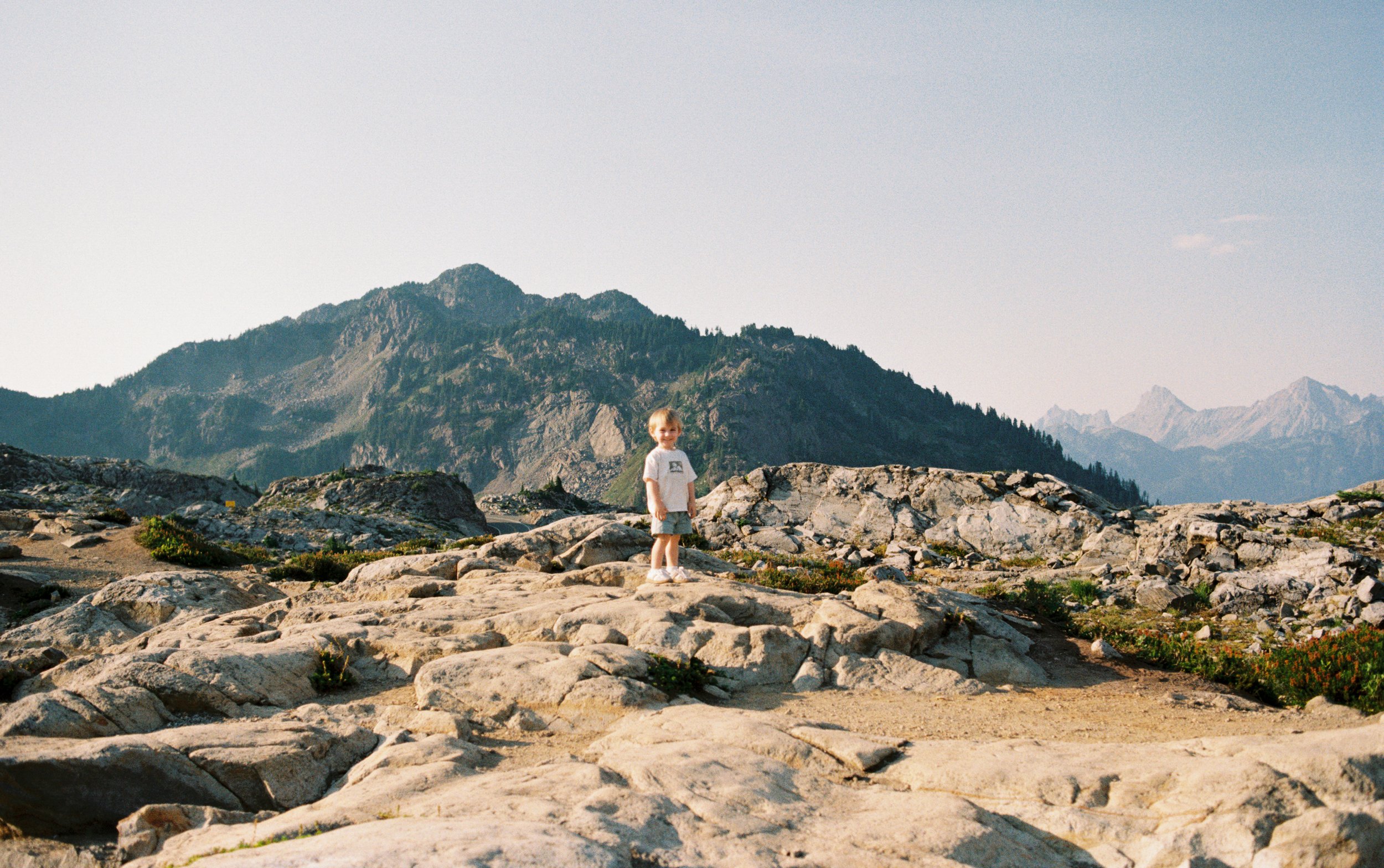 A young child standing on rocky terrain with mountains in the background, under a clear sky.
