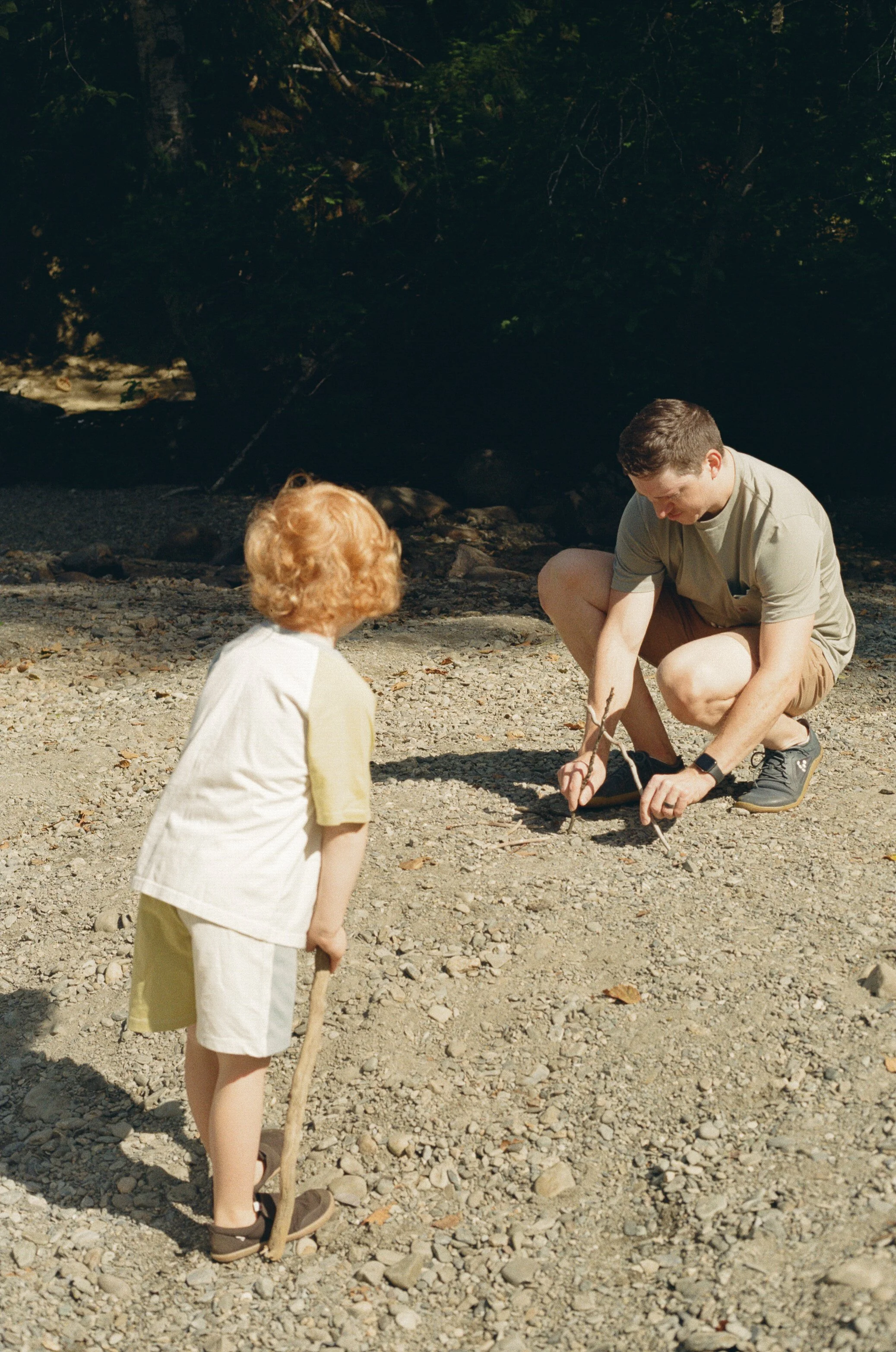 A young man crouching and tying a shoelace on a rocky riverbank while a young girl with curly red hair and light-colored clothing stands nearby, holding a stick. The background shows trees and dark water.
