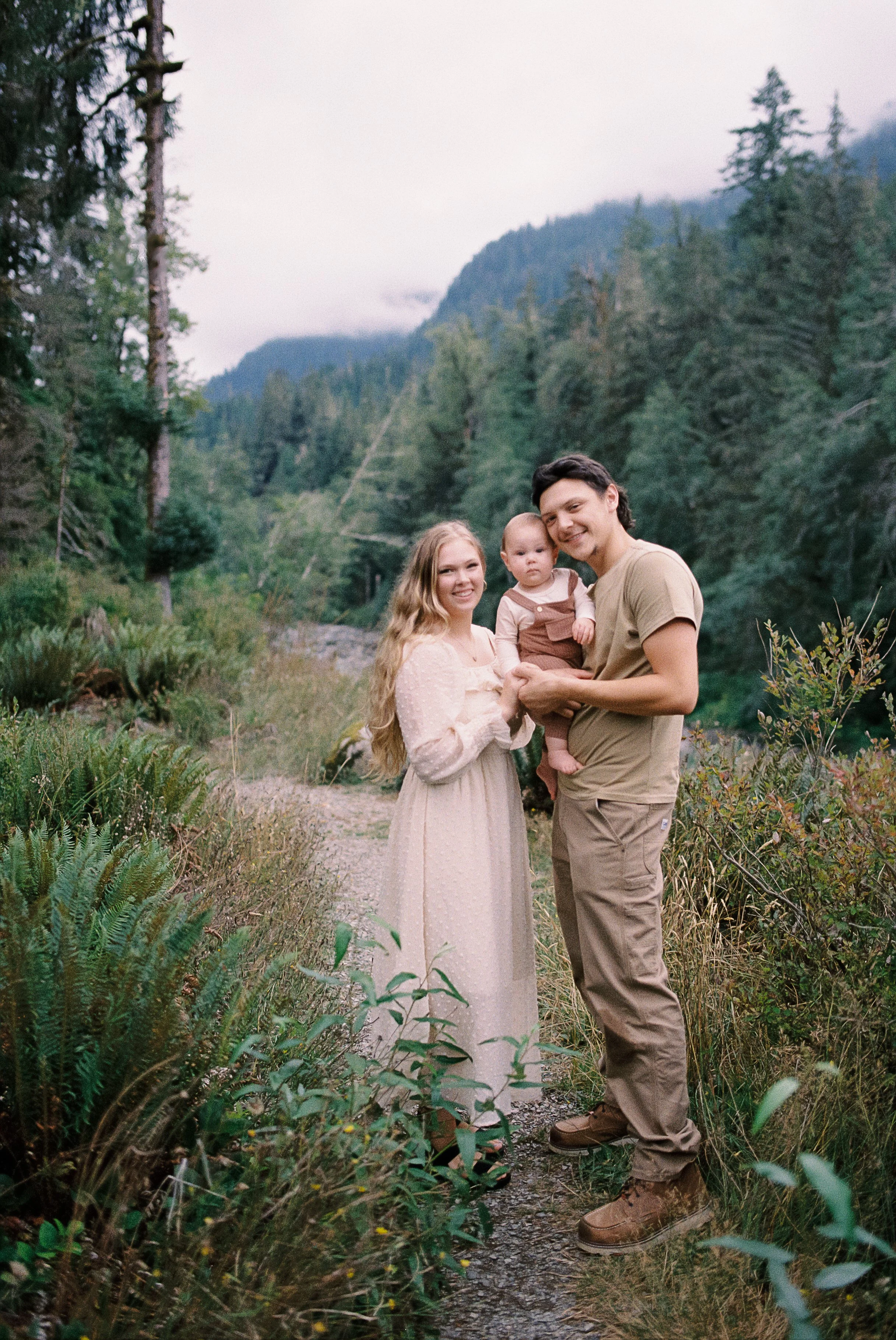 A happy family of three standing on a mountain trail surrounded by lush green trees with mountain scenery in the background.