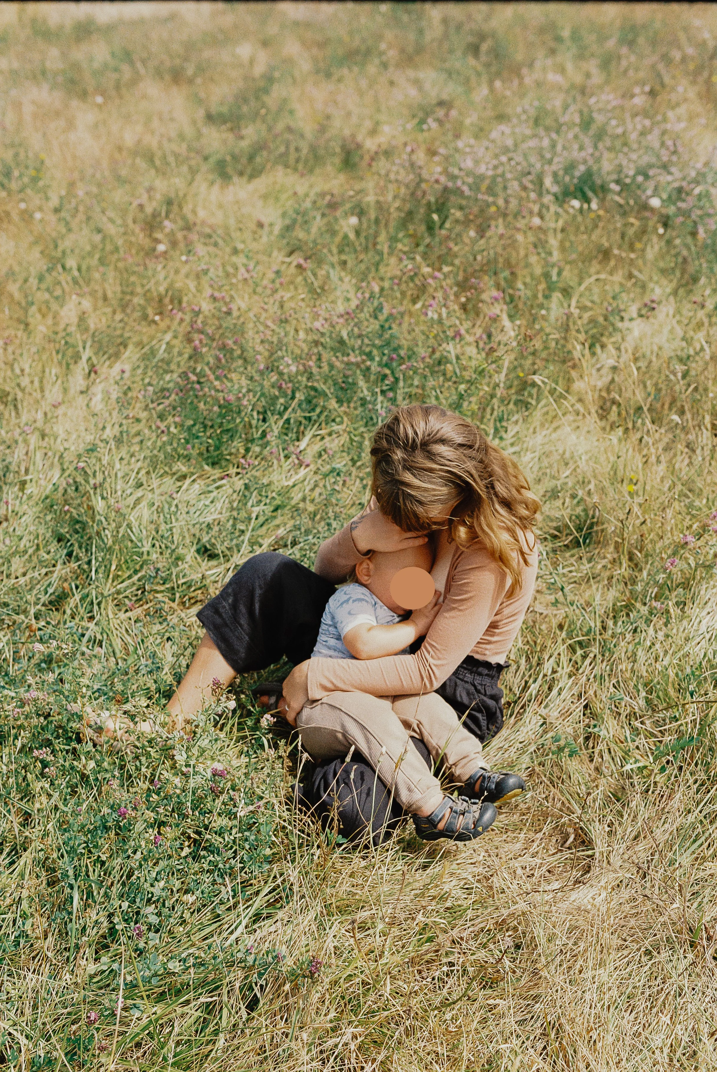 A woman and a small child sitting in a grassy field with wildflowers, the woman is hugging the child close to her chest.