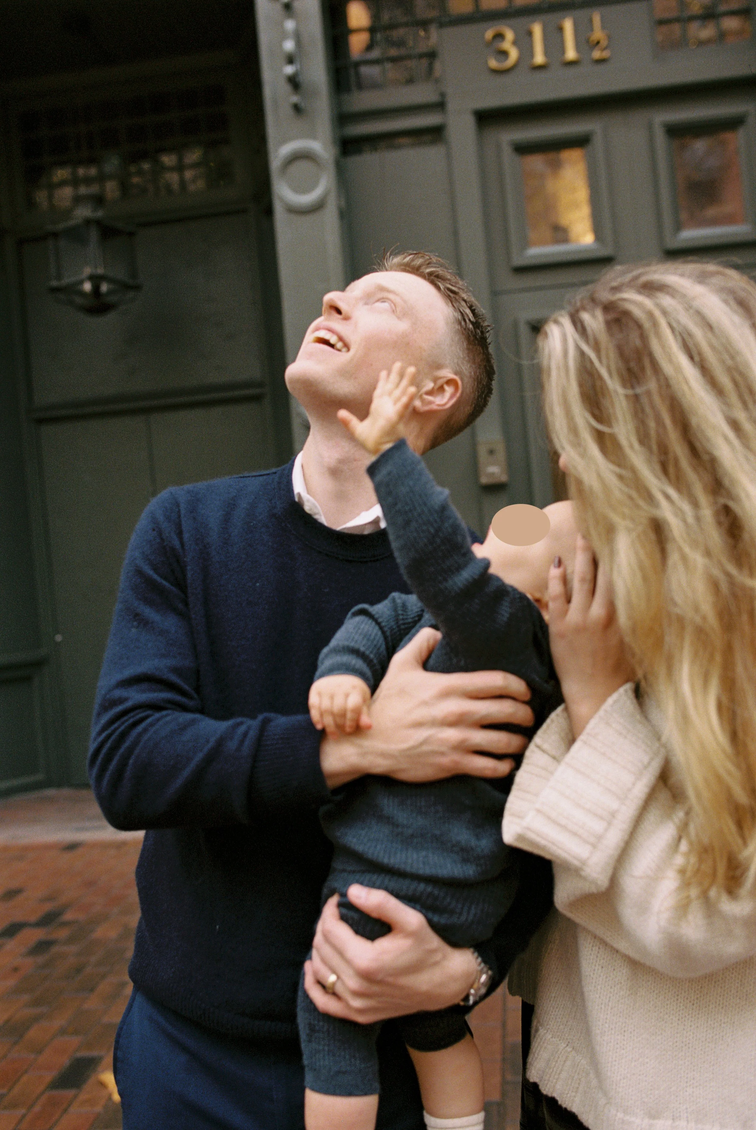A man with short hair wearing a dark sweater is smiling and looking up, while a woman with long blonde hair and a beige coat is touching his face. The man is holding a young child with short hair in his arms.