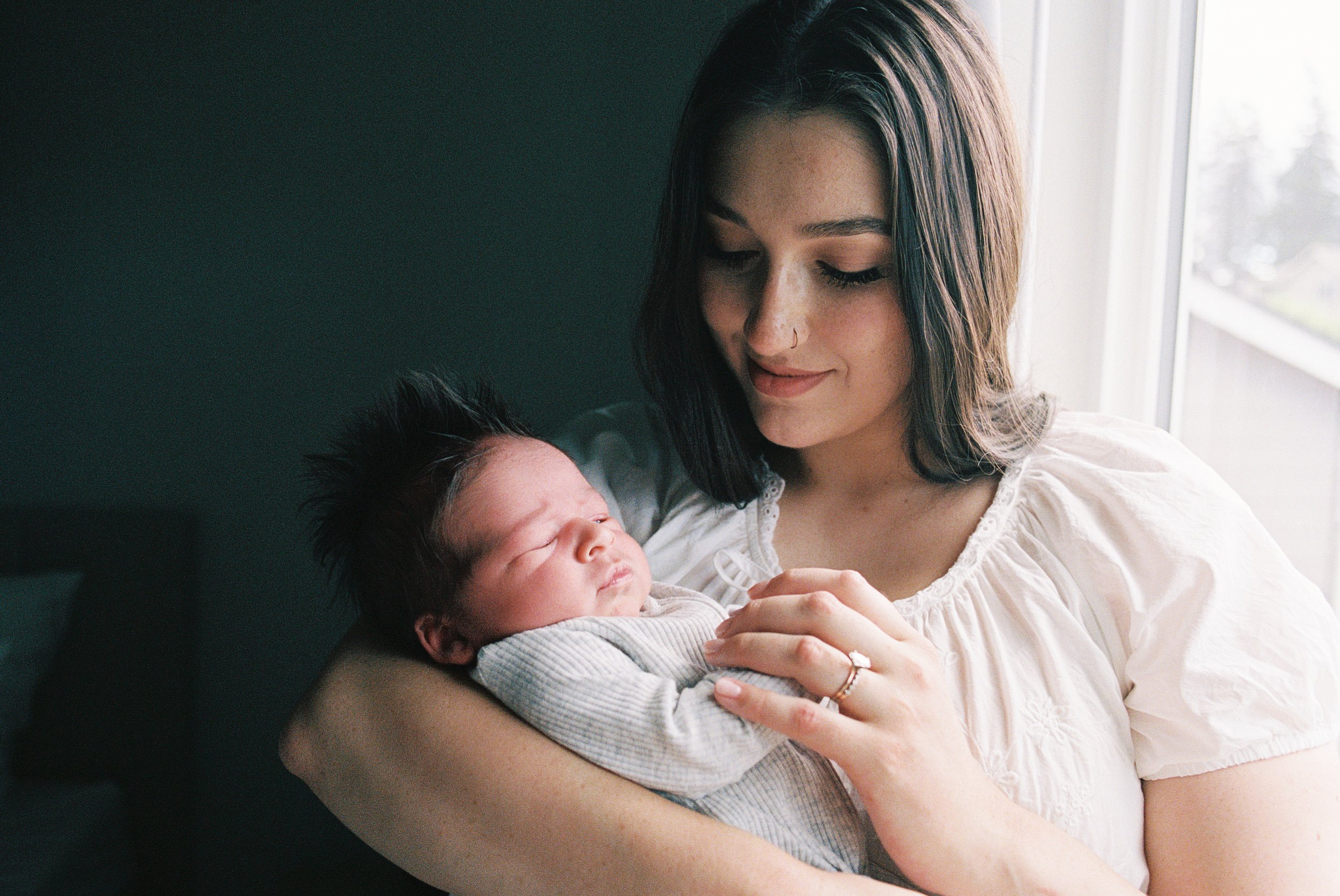 Young woman with dark hair holding a sleeping baby in her arms near a window.