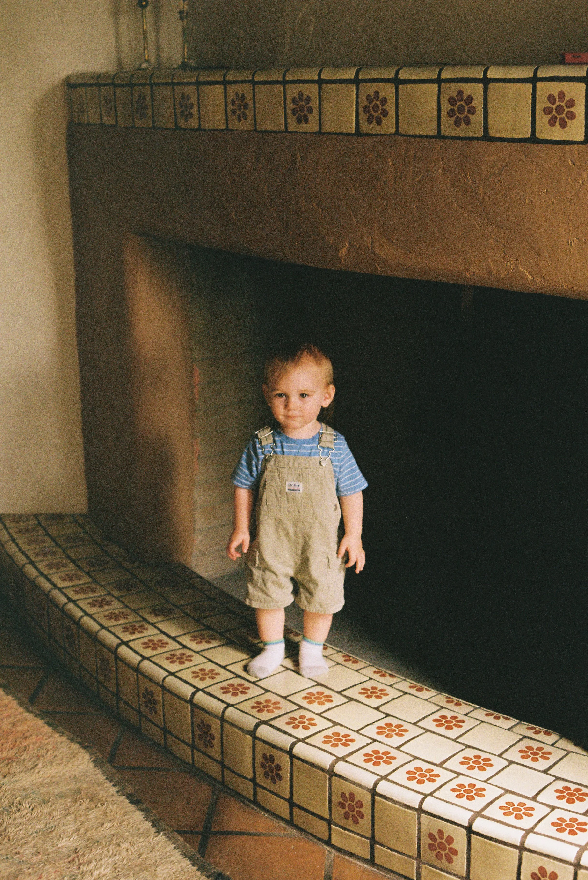 A young child wearing a striped blue shirt, tan overalls, and white socks, standing on a tiled ledge in a room with a fireplace or hearth.