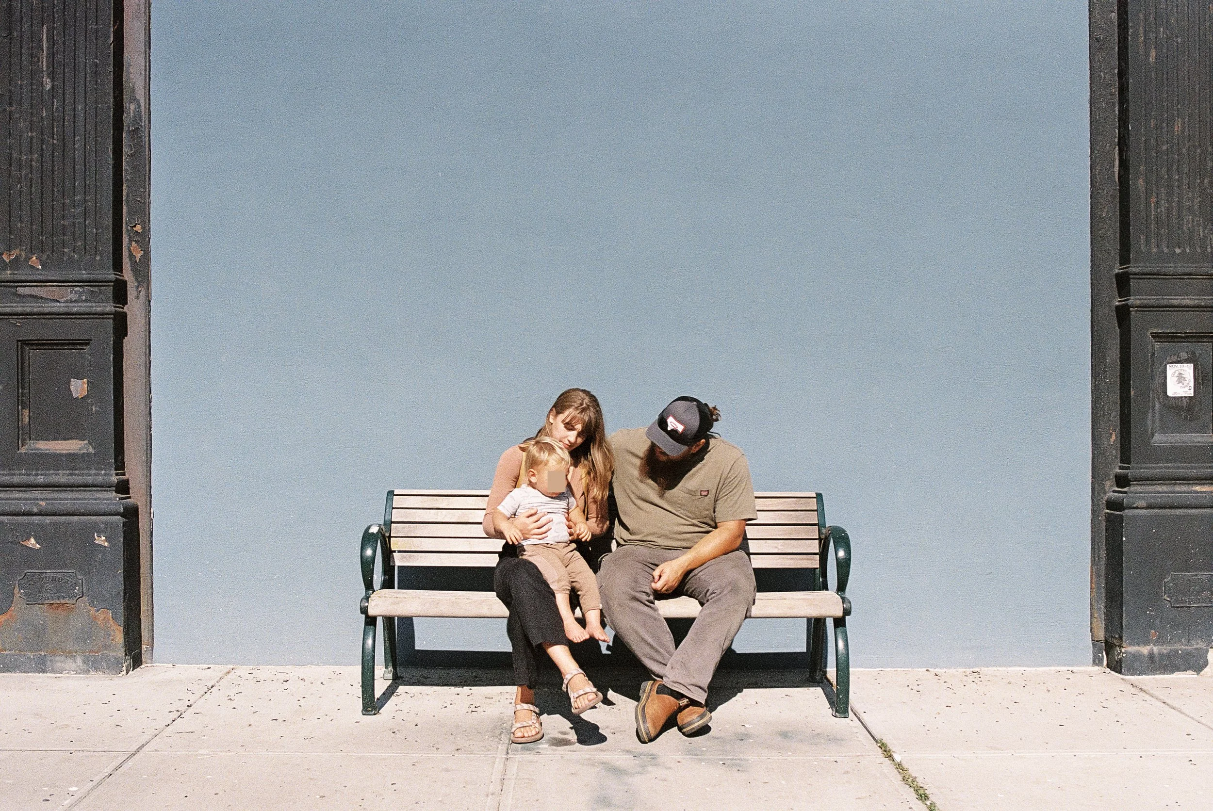 A family of three sitting on a park bench beneath a large open archway, with blue sky visible behind them. The woman holds a young child, and their faces are blurred. The man sits next to them, leaning towards the child. The scene appears warm and re