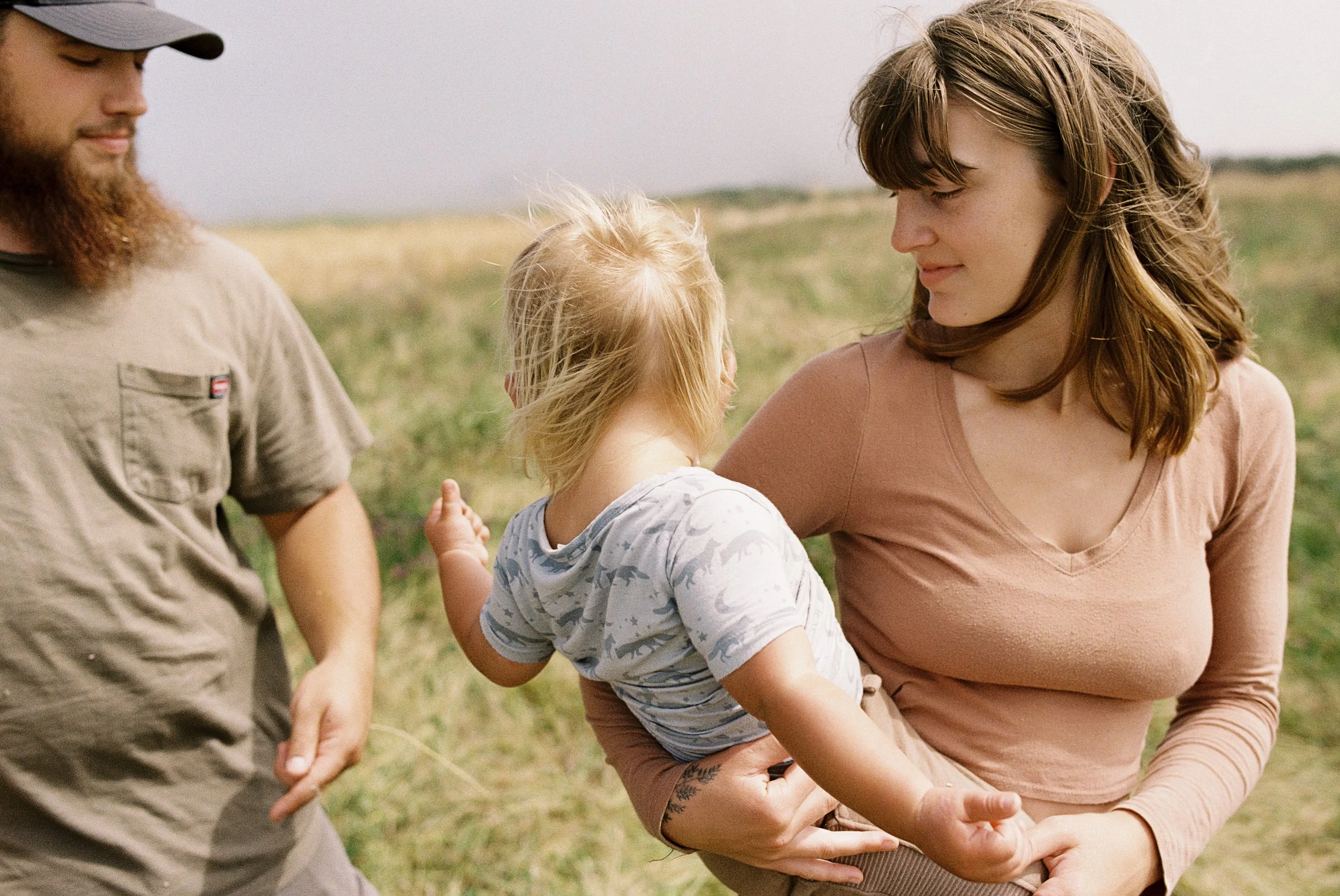 A woman holding a young child in an outdoor grassy area, with a man standing nearby. The scene appears peaceful and intimate, with a blurred background of open land.