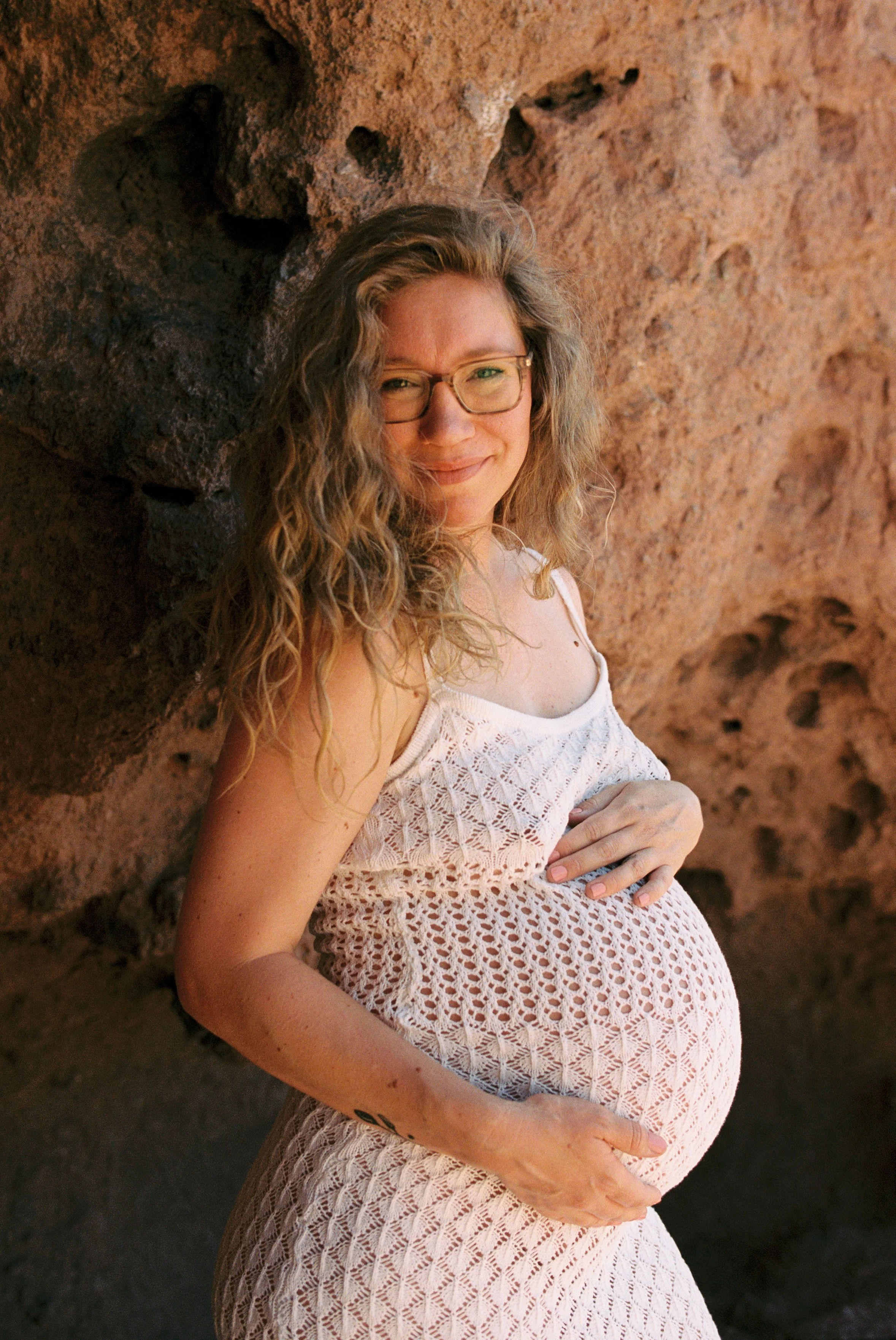 A pregnant woman with curly hair and glasses standing outdoors against a rocky background, smiling and gently cradling her pregnant belly with her hands.