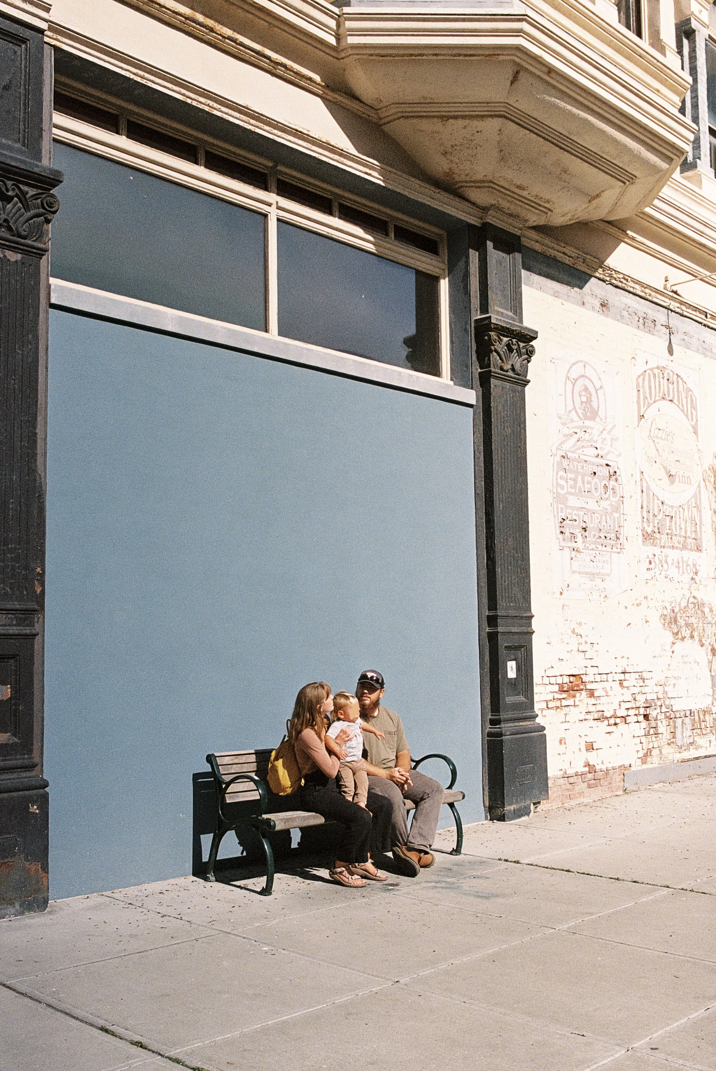 A family of three sitting on a bench outside a building with a brick and painted wall. The woman holds a young child, and the man sits beside them.