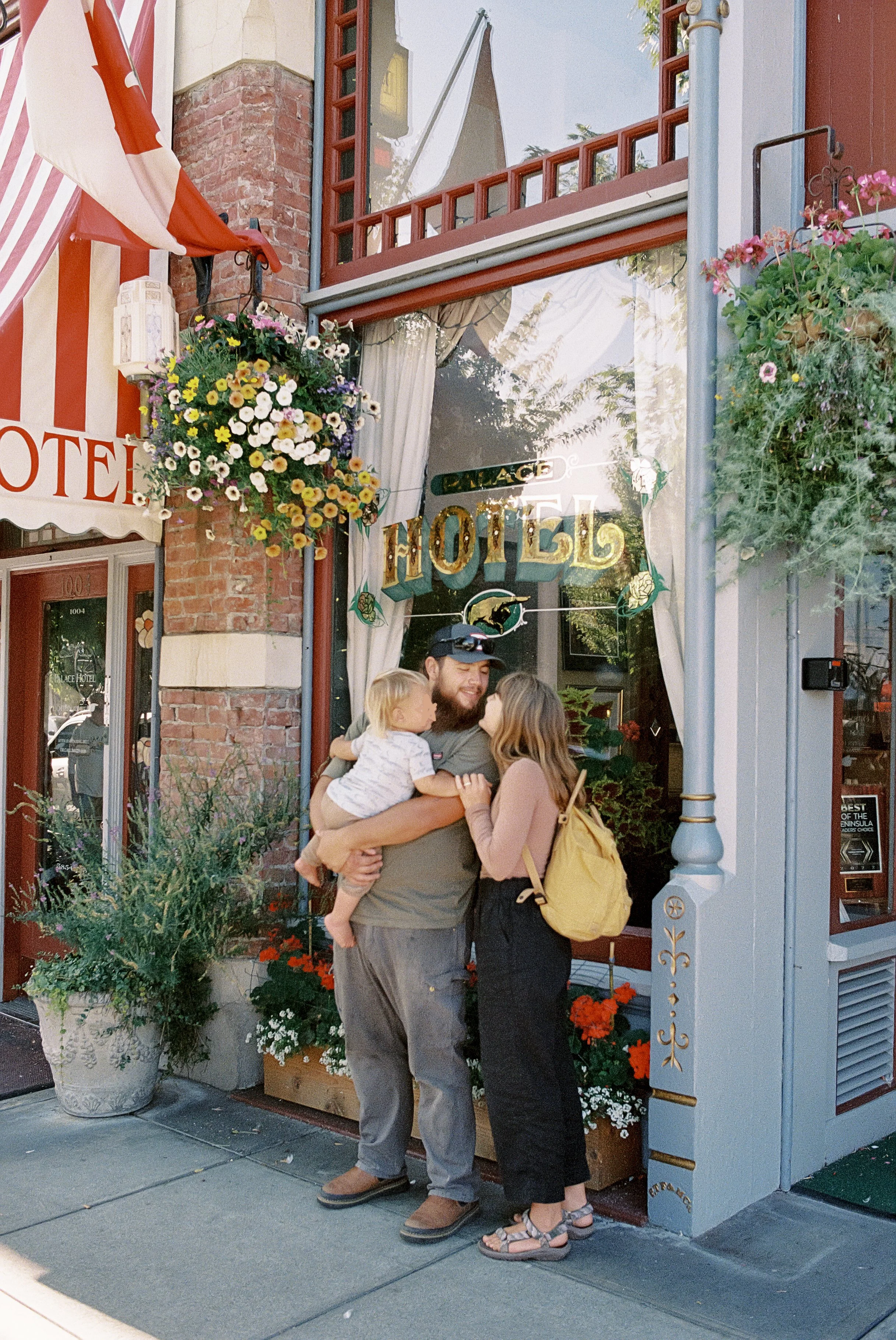 A man holding a child and a woman standing in front of a colorful storefront with floral decorations and a gold sign that reads 'hotel'.