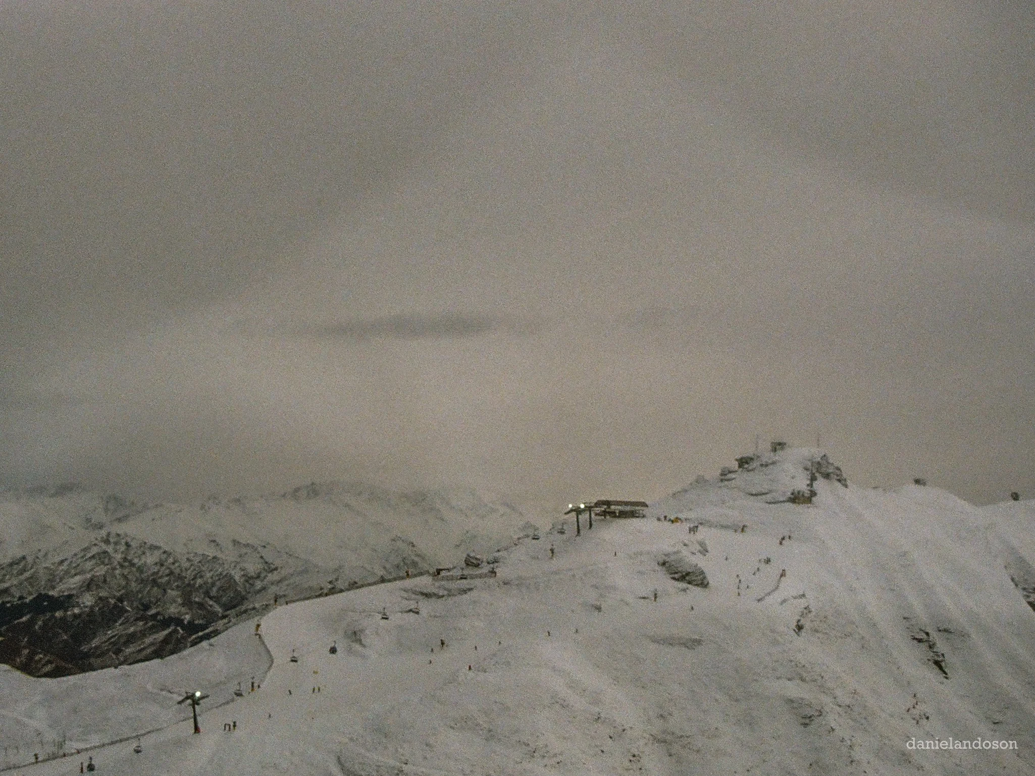 Snow-covered mountain landscape at night with ski path, ski lift, and ski poles in the foreground.