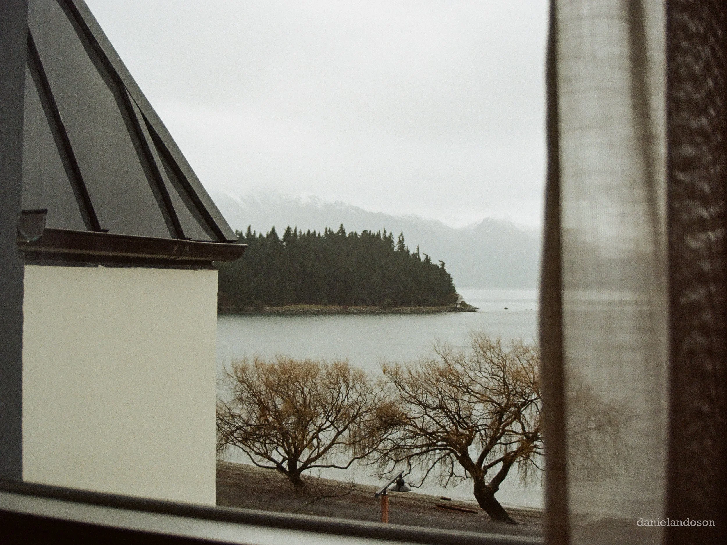 A view of a lake or river with tree-covered hills in the distance, seen through a window with partially drawn curtains, during overcast or rainy weather.