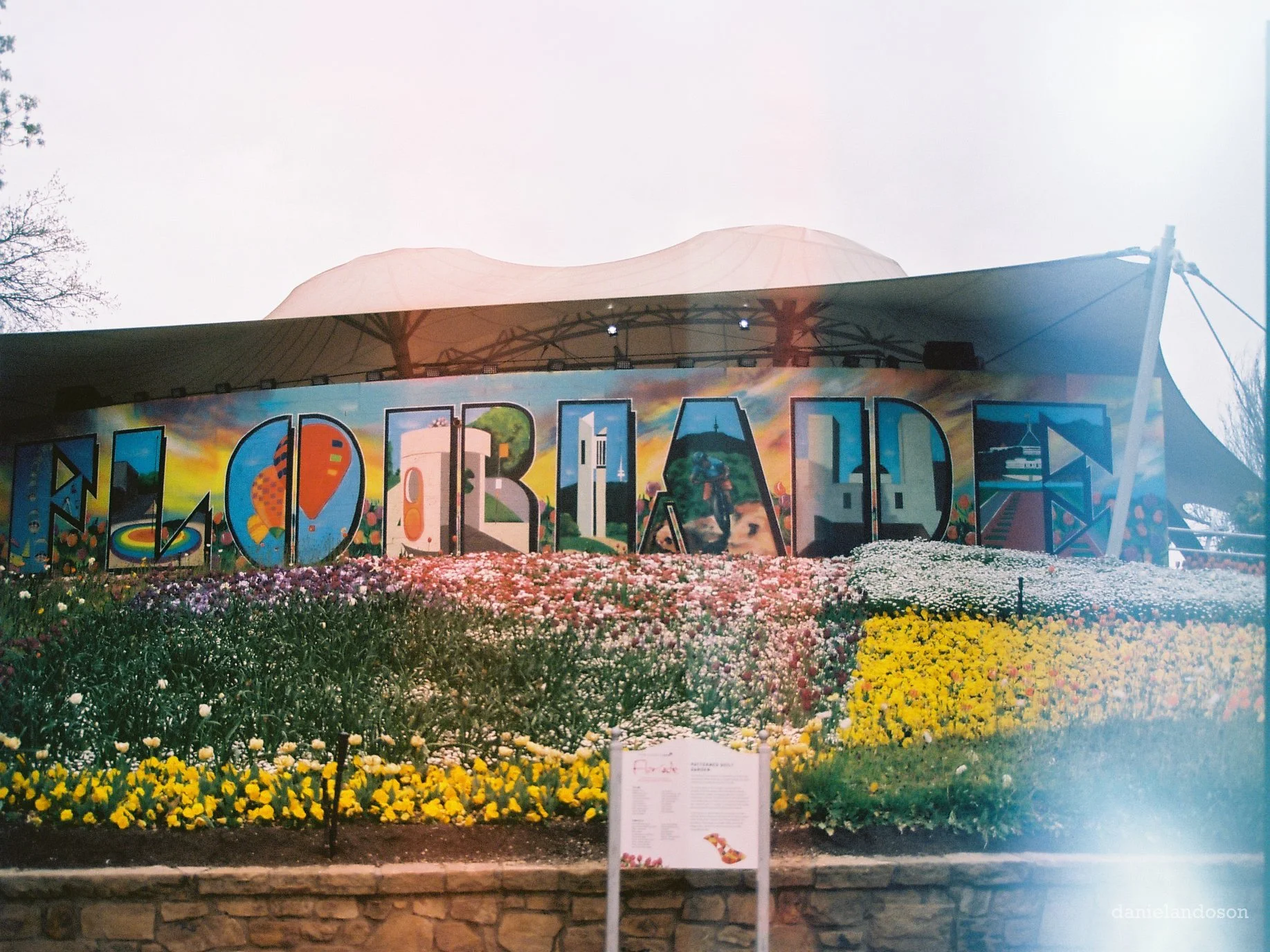 A colorful mural spelling out "HELLOA" with a vibrant garden of pink, white, yellow, and purple flowers in the foreground and a white canopy tent above.