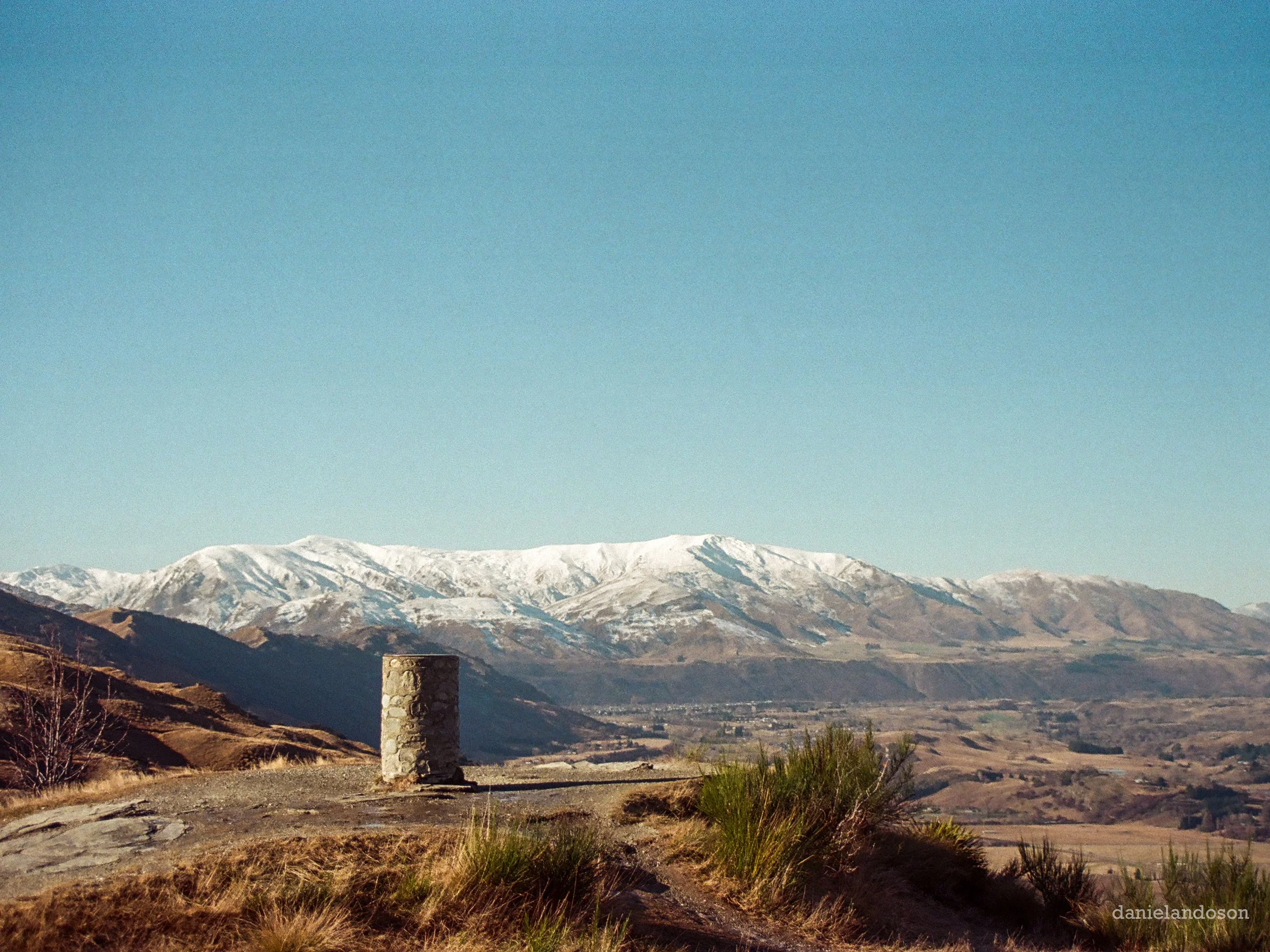 Mountains with snow caps under a clear blue sky, seen from a foreground with dry grass and a stone pillar.