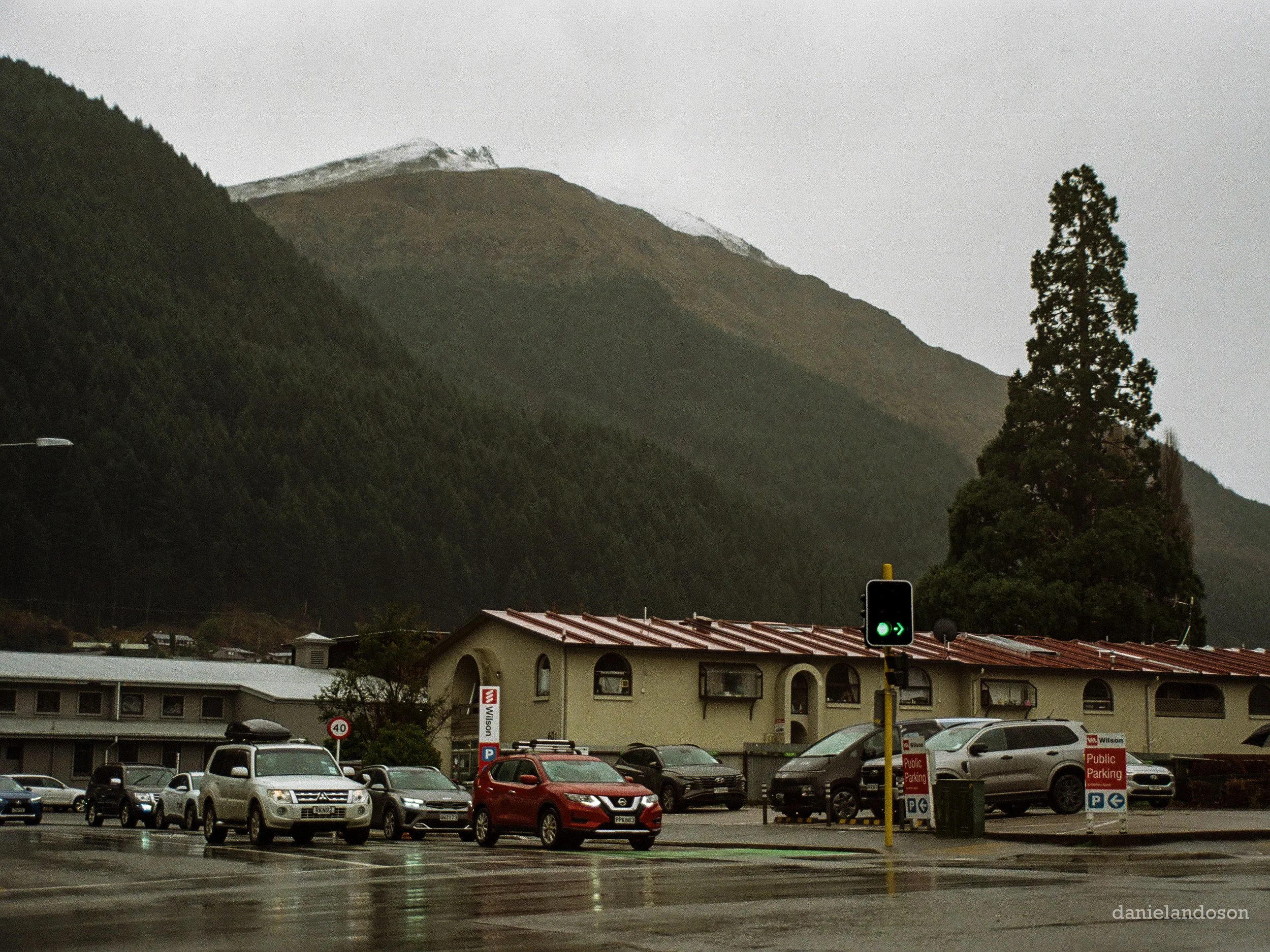 Parking lot with several cars in front of a low building, hillside with snow-capped mountains in the background, and a traffic light showing green arrow.