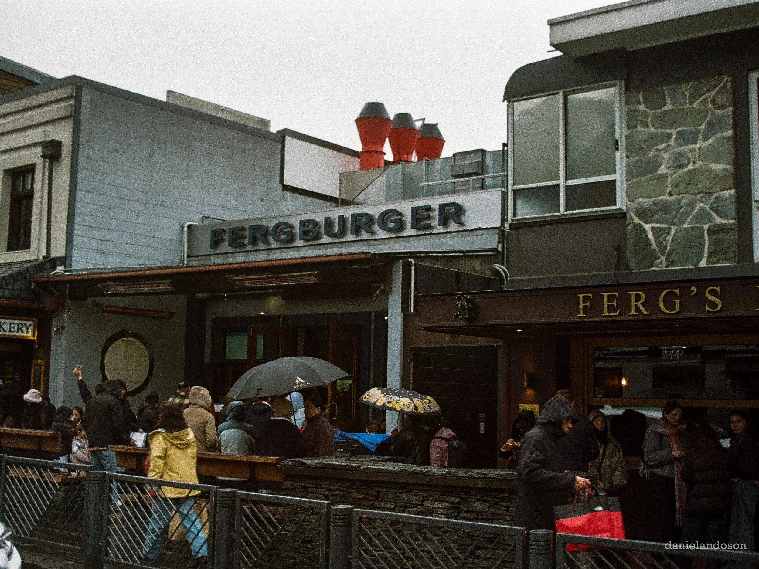 People gathered outside a restaurant named Fergburger, with umbrellas, under a cloudy sky.