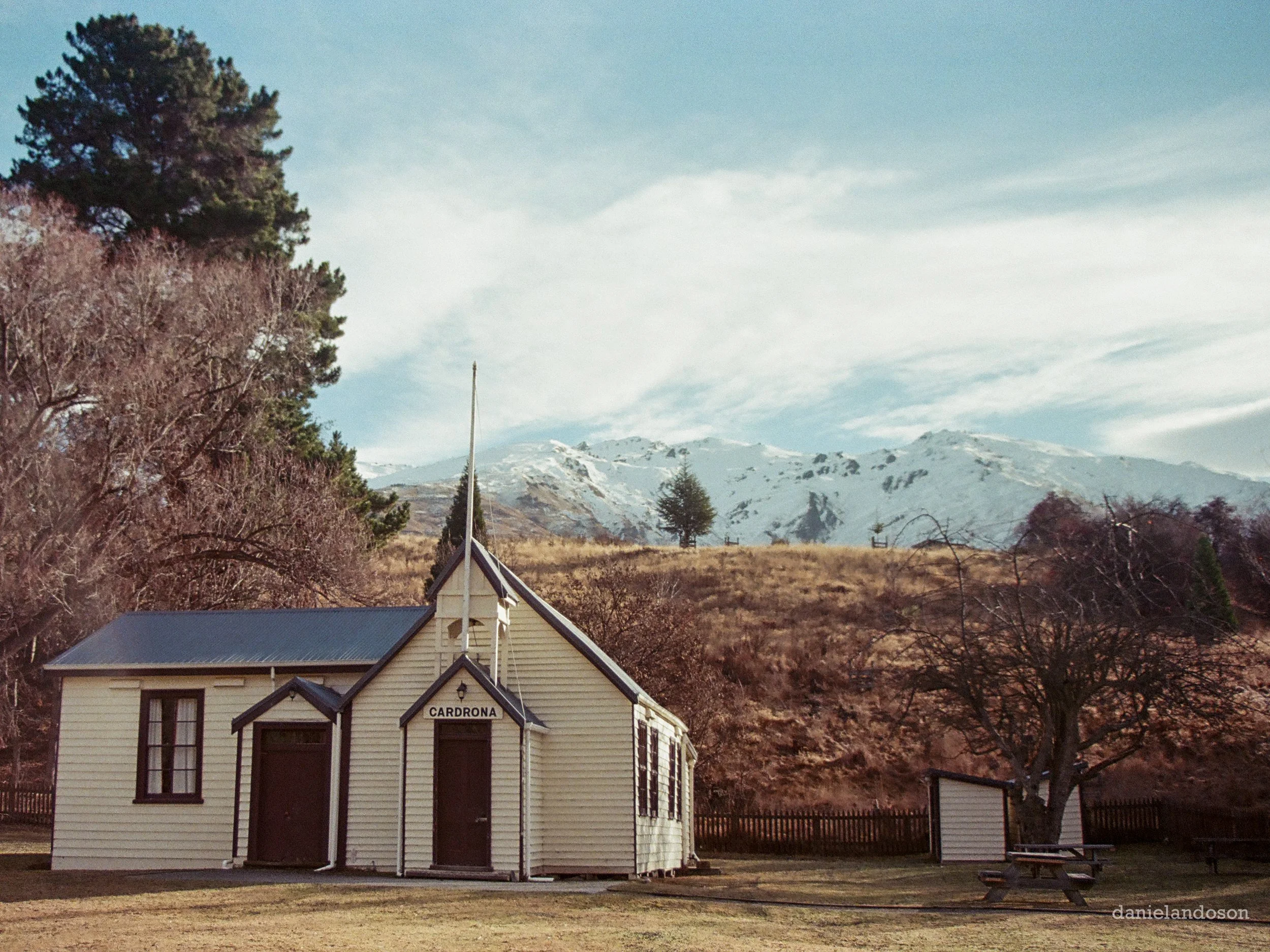 A vintage-style yellow building with black trim, gabled roof, and a sign that says 'Cardrona,' situated in a rural landscape with mountains in the background, trees in the foreground, and the sky overhead.
