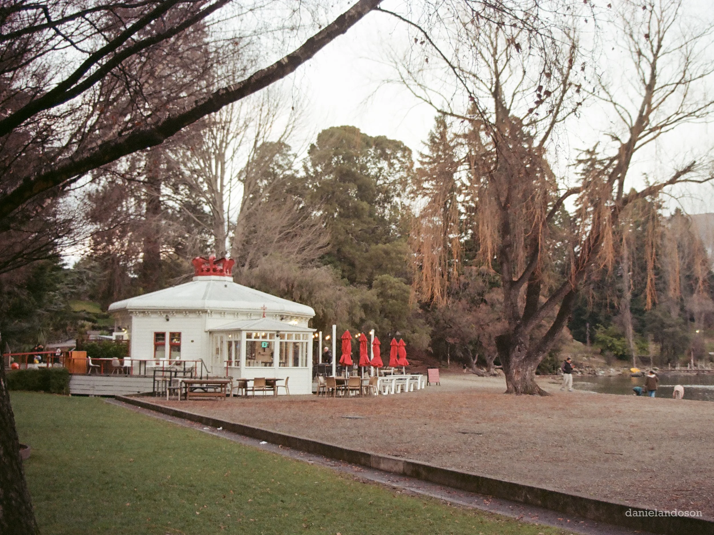 A park scene with a white pavilion with a red roof, surrounded by trees, some with bare branches and others with dried hanging leaves, and red umbrellas with chairs underneath, on a cloudy day.