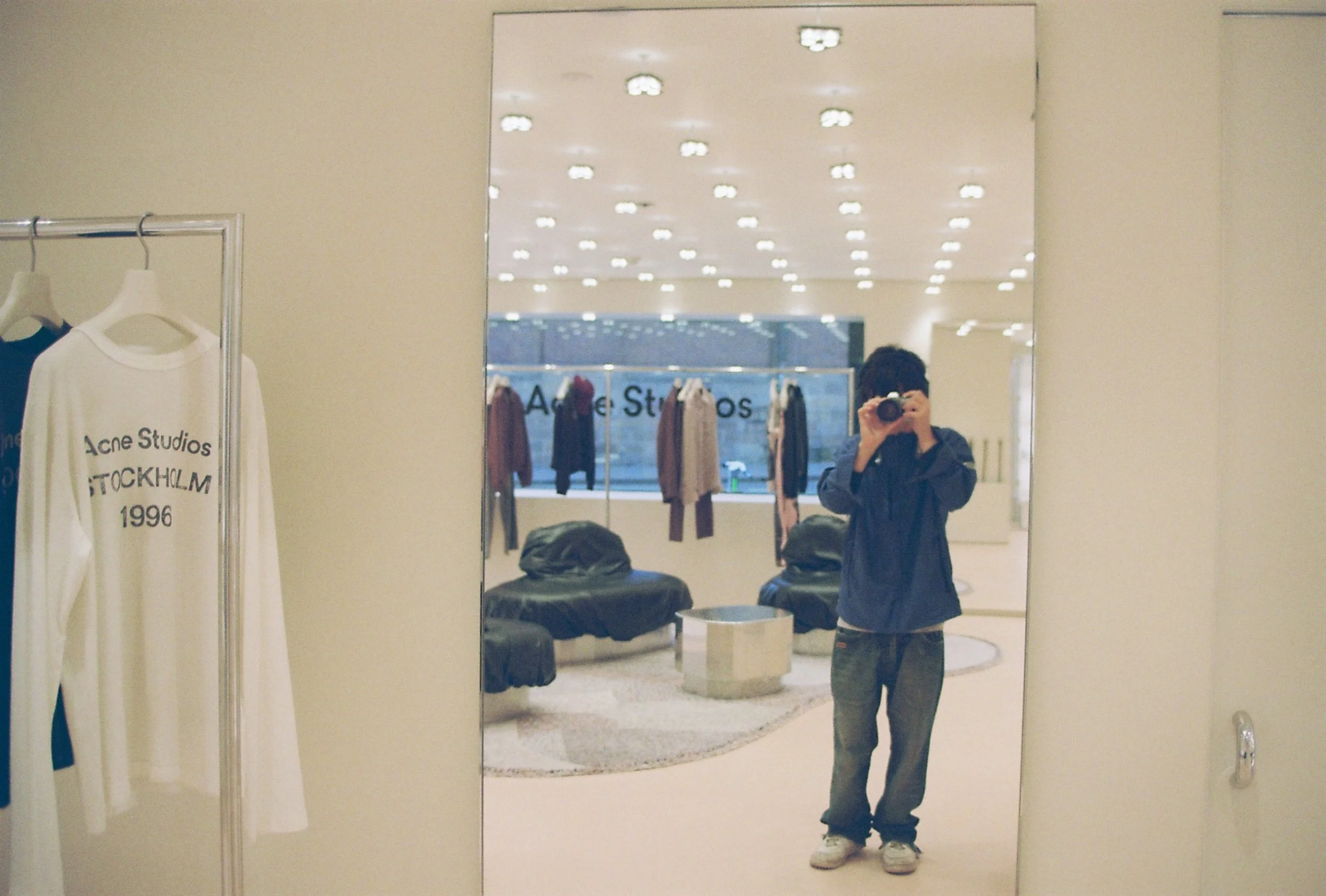 A person taking a mirror selfie in a store fitting room, wearing a dark jacket, gray pants, and white shoes. There are clothing racks and shopping bags visible in the background.