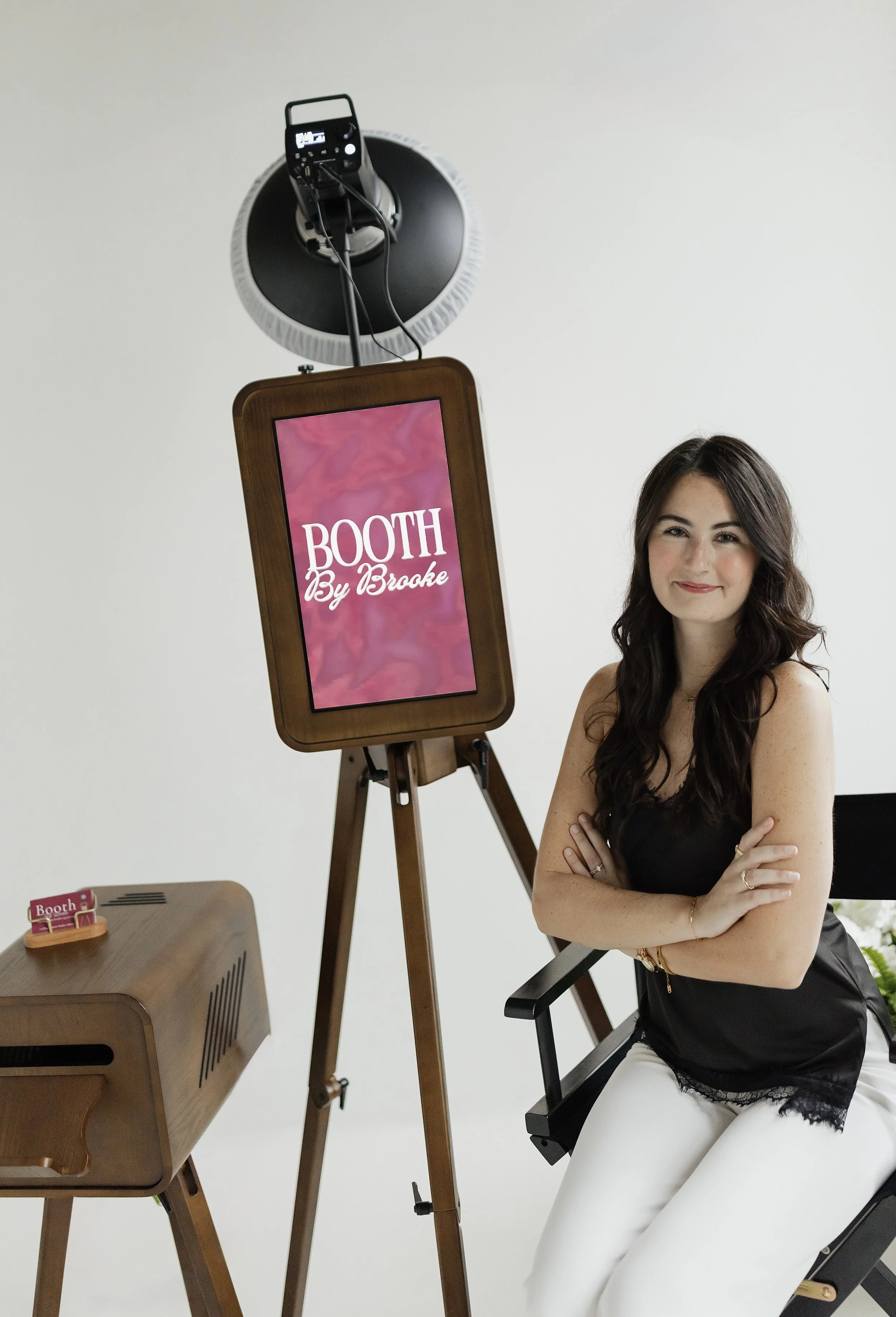 A woman with dark hair and a black top sitting on a director's chair next to a photo booth sign, with studio equipment in the background.