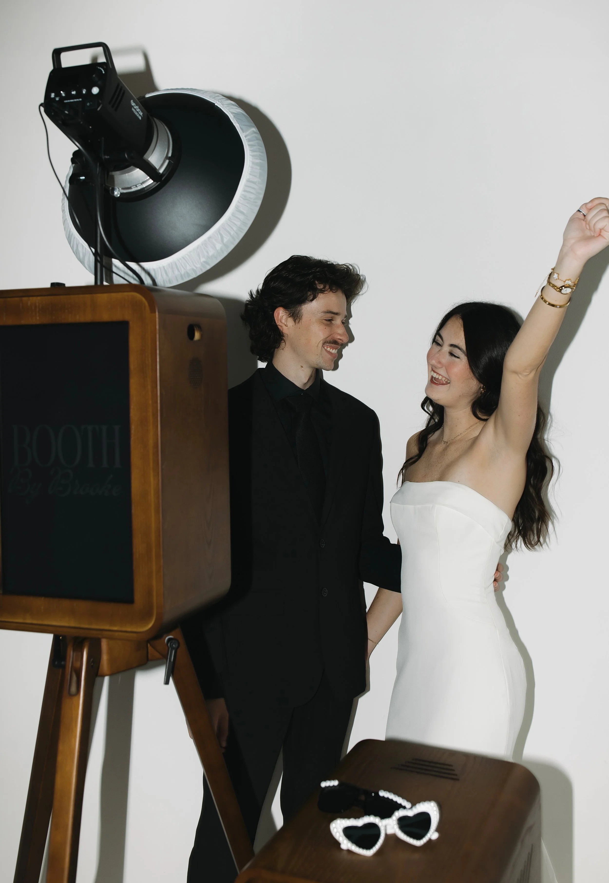 A woman in a white strapless dress and a man in a black suit are smiling and looking at each other, with the woman raising her right arm, in a photo shoot setting with studio lighting. There is a vintage wooden photo booth in the foreground, and a pair of heart-shaped sunglasses on a wooden table.