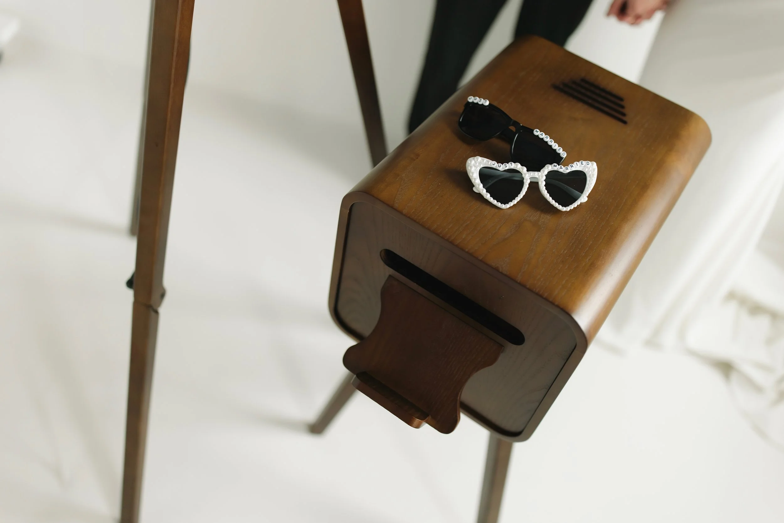 On a wooden chair, there are a pair of black sunglasses with white beads spelling "GOD MARRY ME" and another pair of white beaded sunglasses with the words "JUST MARRIED" on the frames.